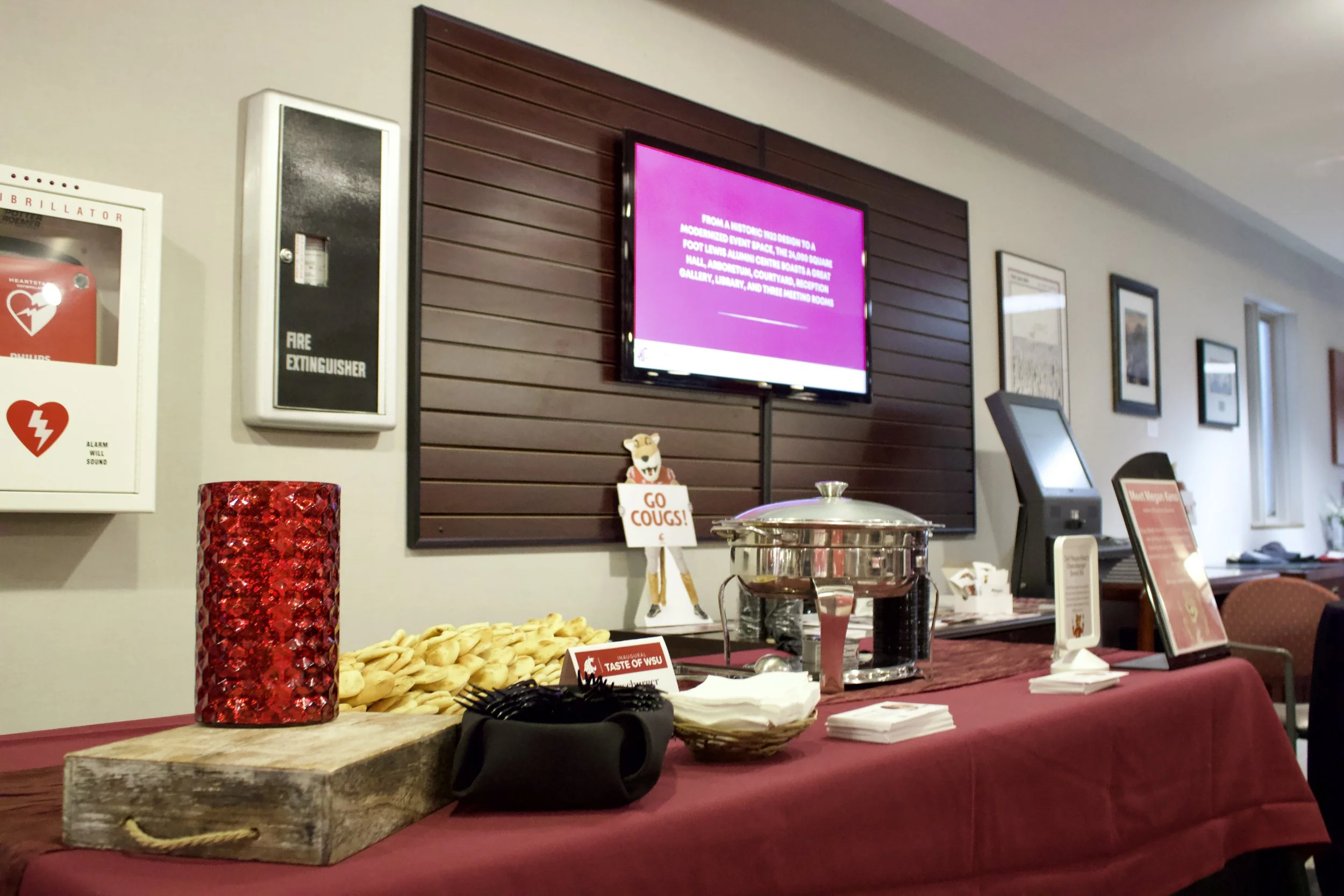Buffet table with cookies, a red mosaic vase, and sign that says 'Go Cougs!' in a room with framed pictures, a TV, and electronic equipment in the background.