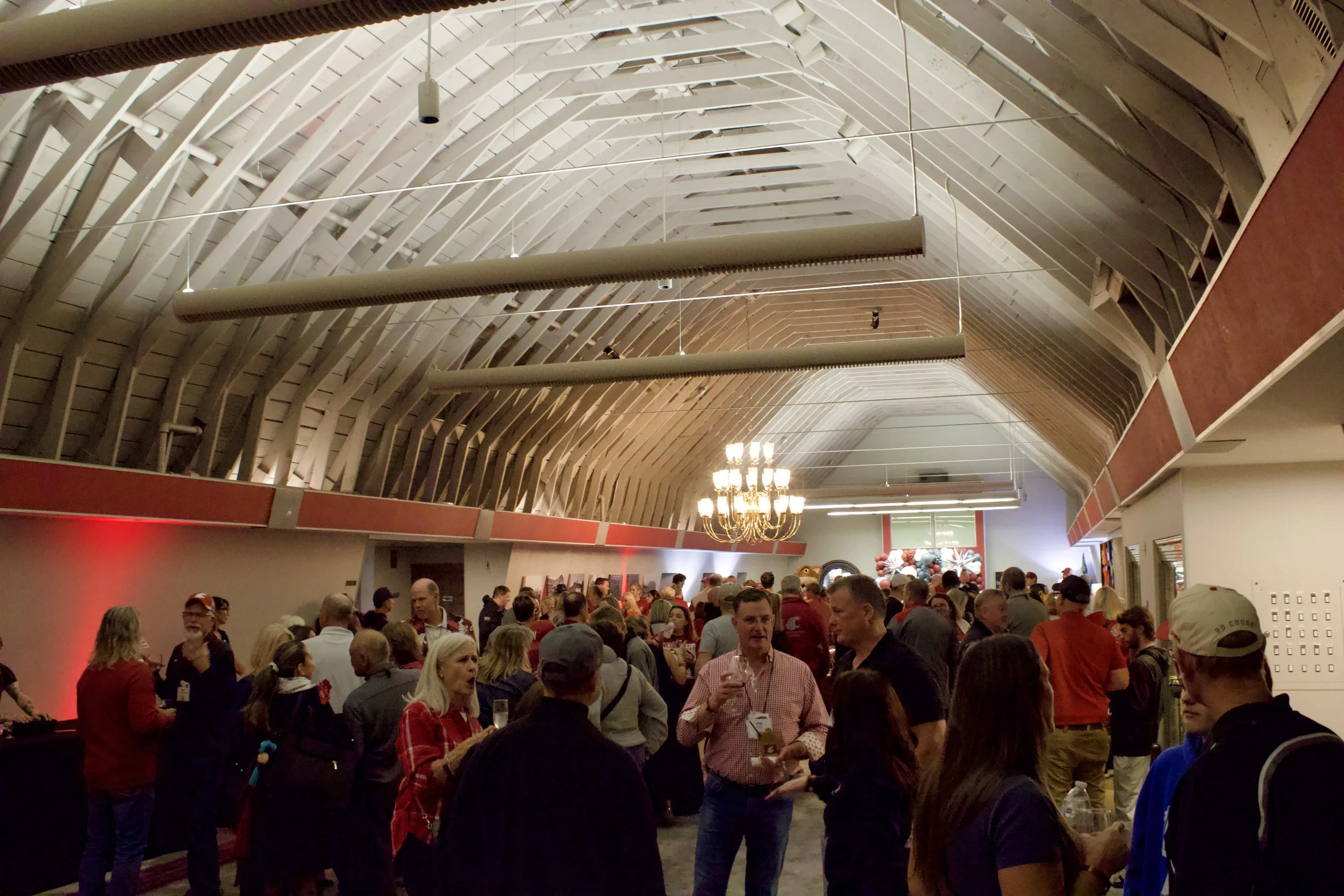 A large indoor event with many people gathered, standing and socializing, in a room with a high vaulted wooden ceiling and a chandelier.