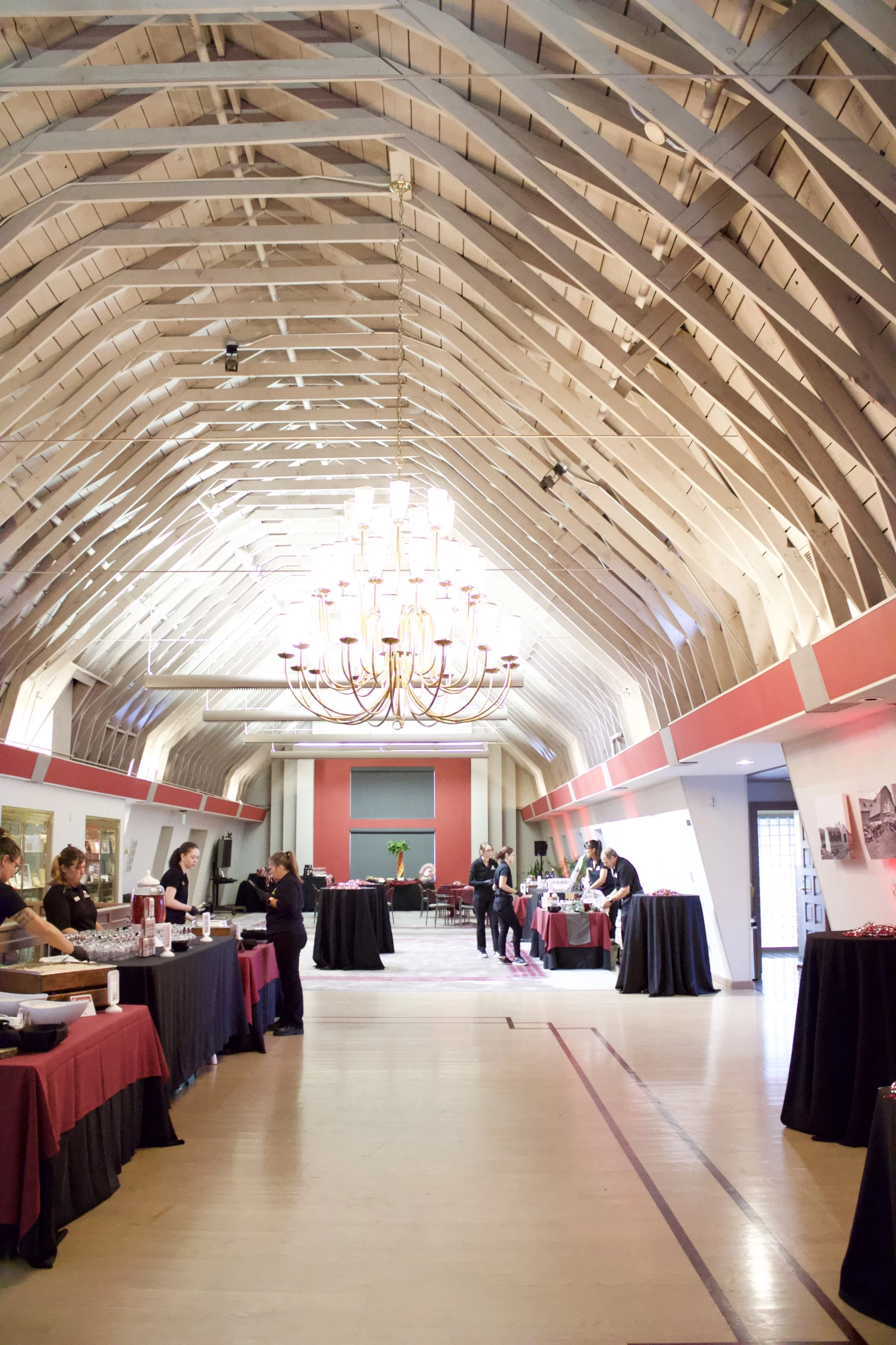Indoor event space with high arched wooden ceiling, large chandelier hanging in the center, and tables with black and red tablecloths being set up by staff.
