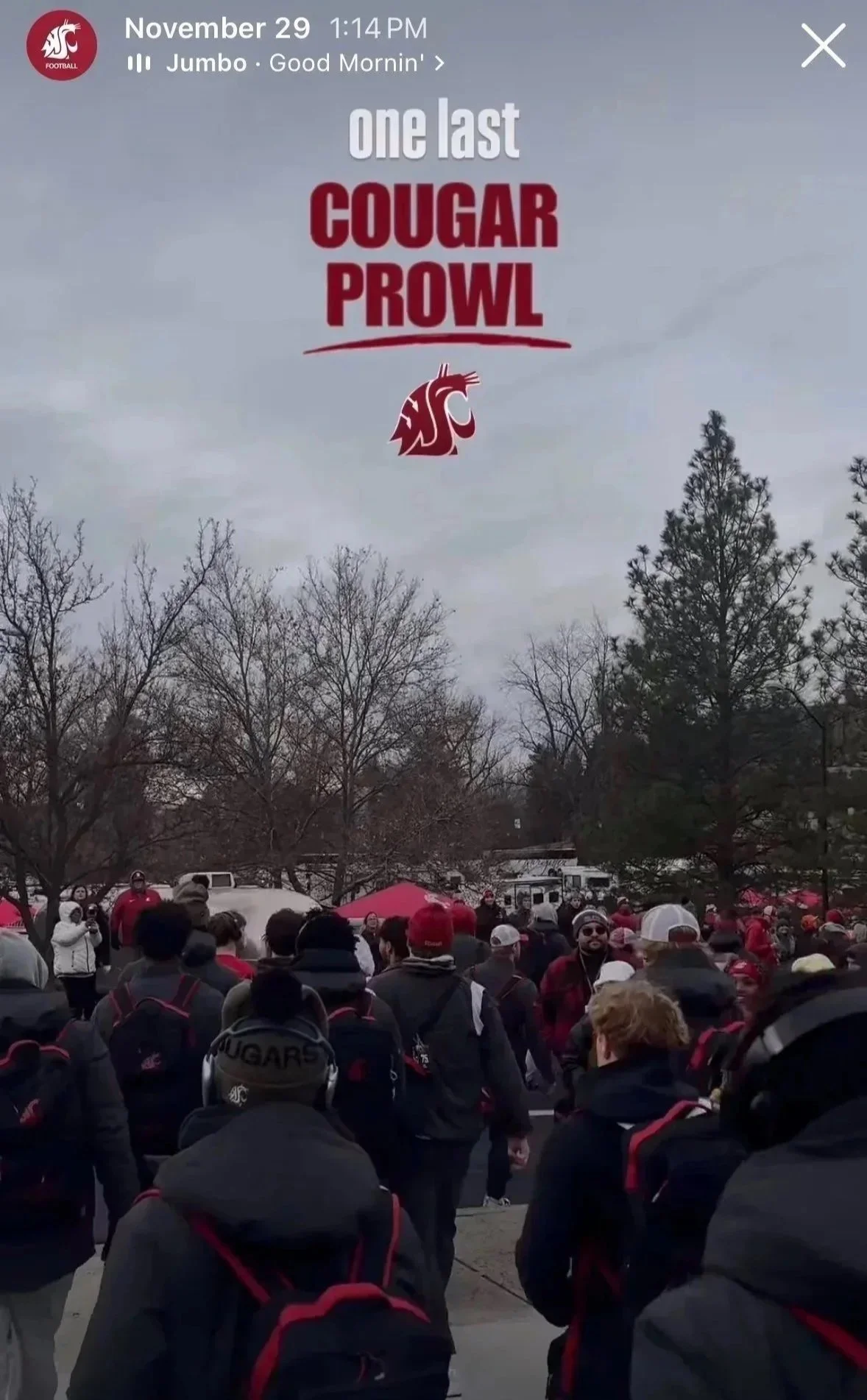 A crowd of people gathered outdoors on a cloudy day, many wearing red and black clothing, at what appears to be a university event or rally, with trees without leaves in the background.