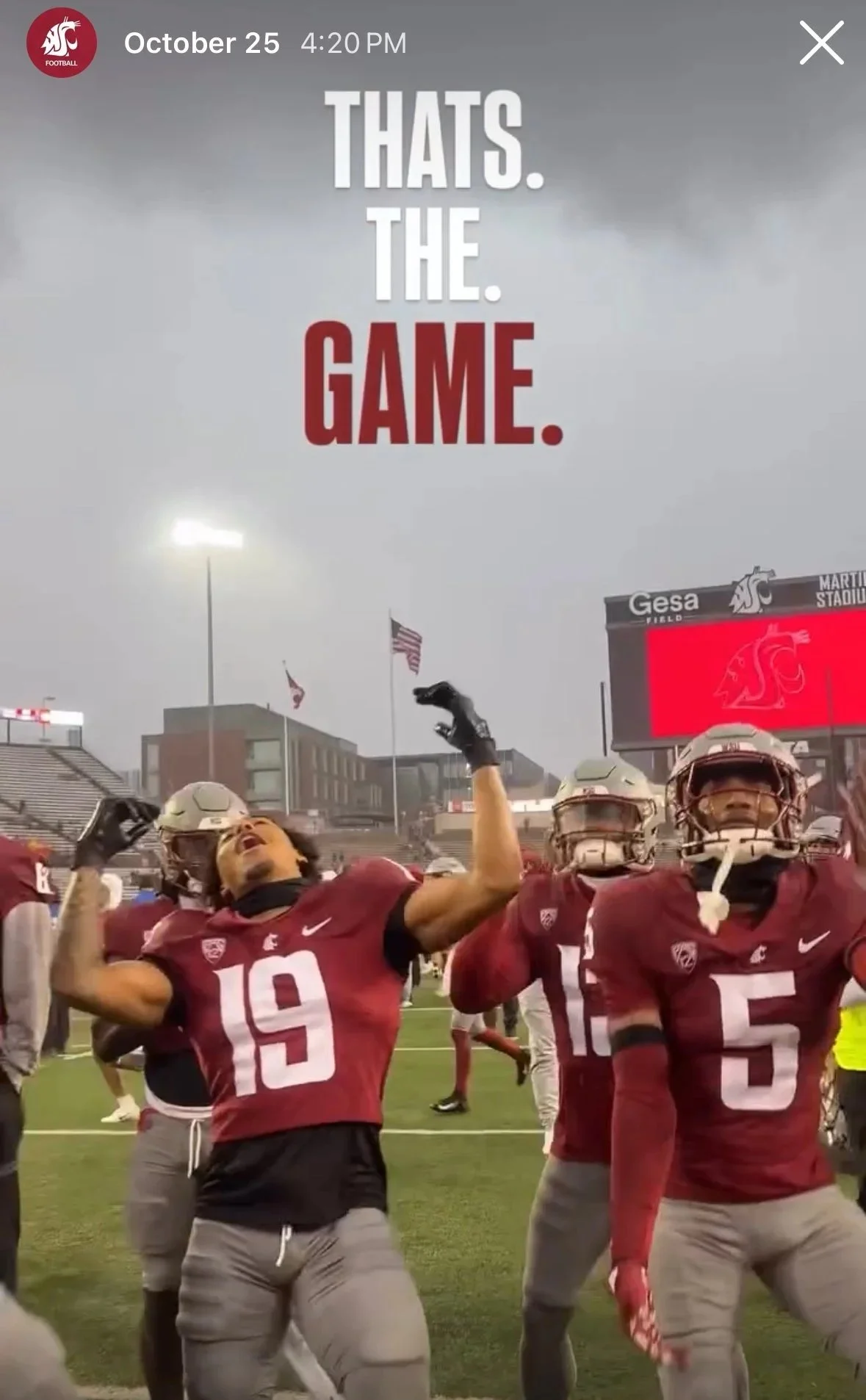 Football players in red jerseys are celebrating on the field at a stadium, with a large digital scoreboard in the background displaying the team's logo, and an overcast sky overhead.