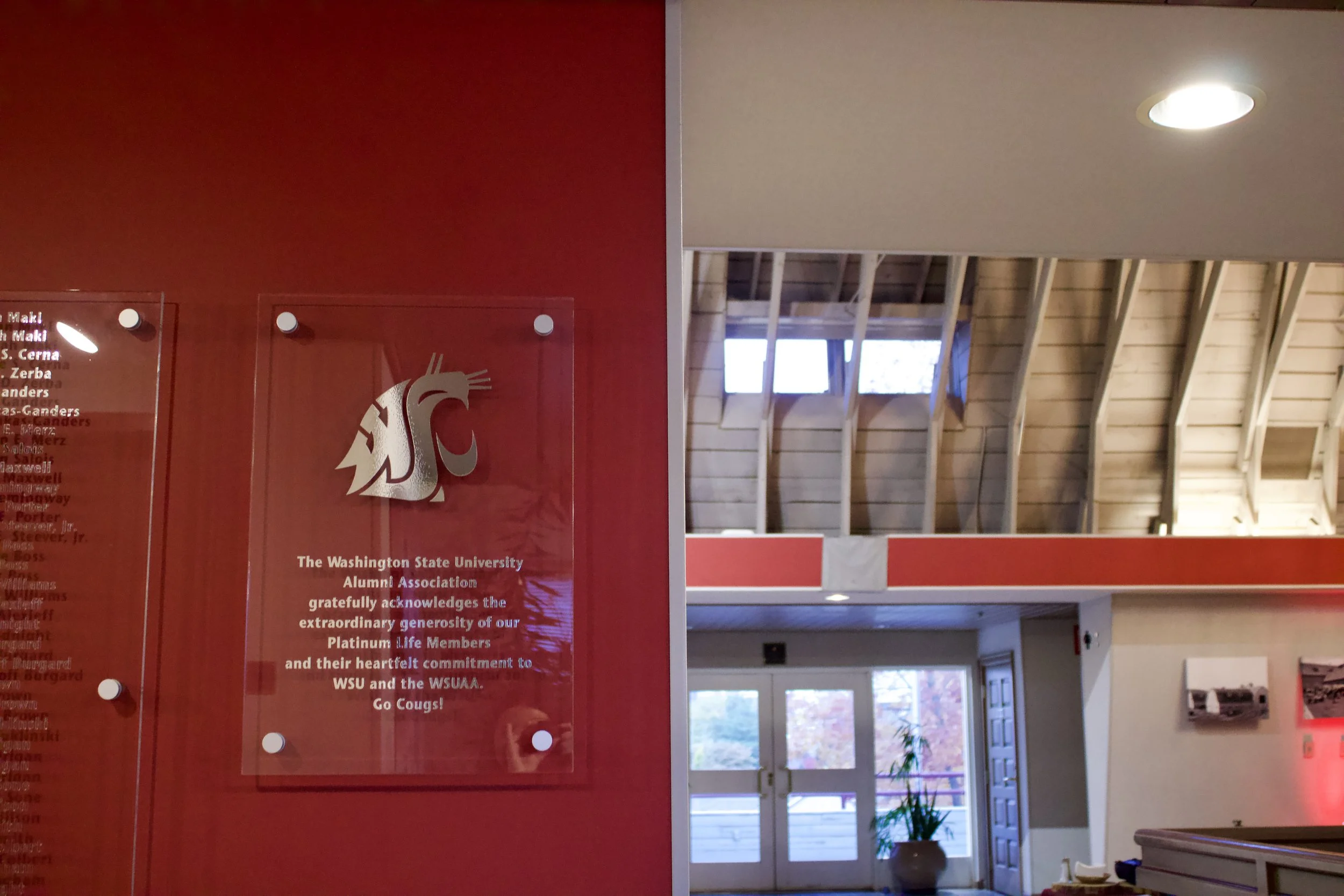 Plaque with Washington State University logo on a red wall, acknowledging alumni donations. Modern interior building with wooden ceiling, skylights, and glass doors leading outside.