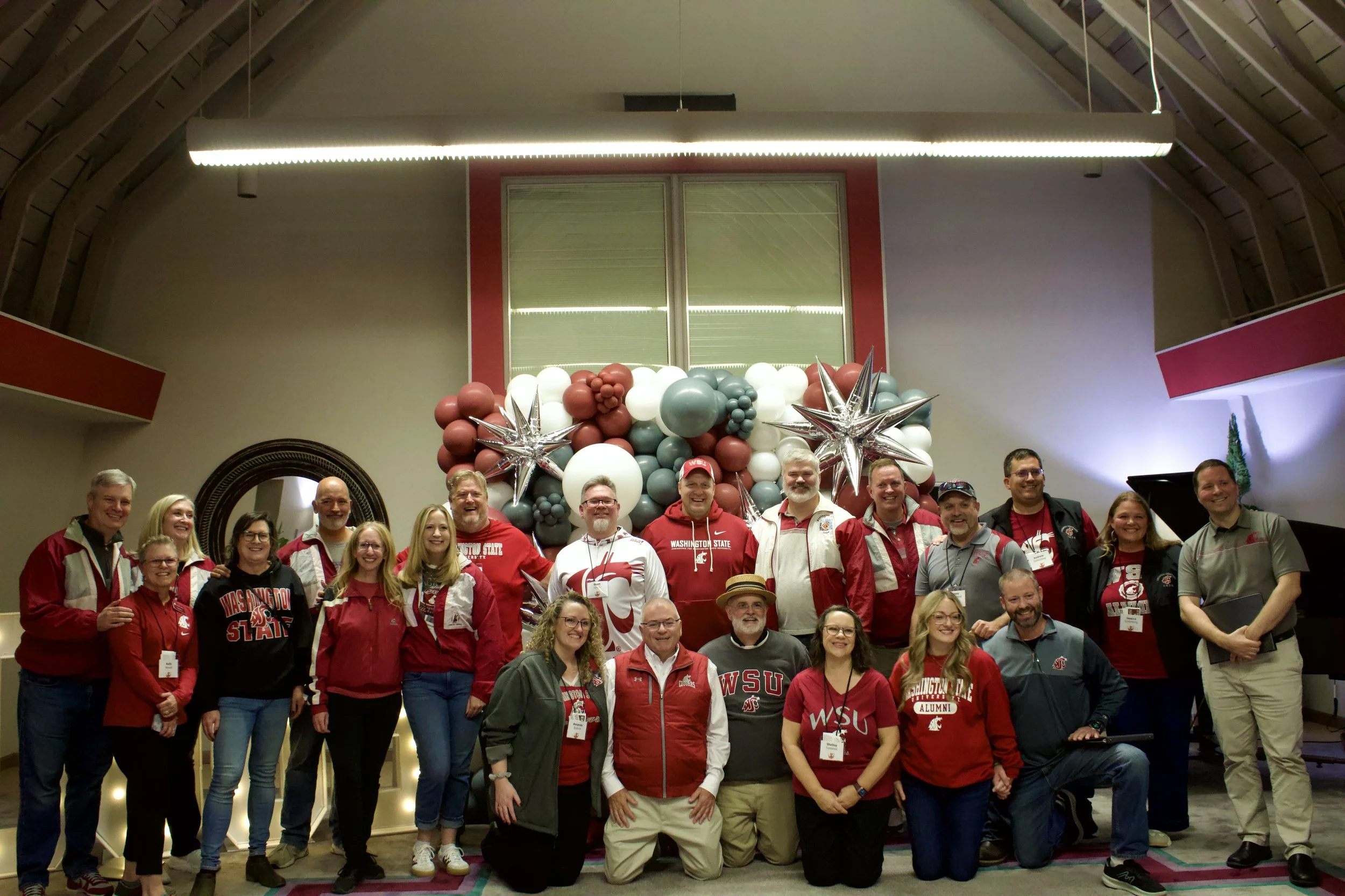 Group of people wearing Washington State University apparel, standing in front of a balloon decoration with stars, in an indoor venue.