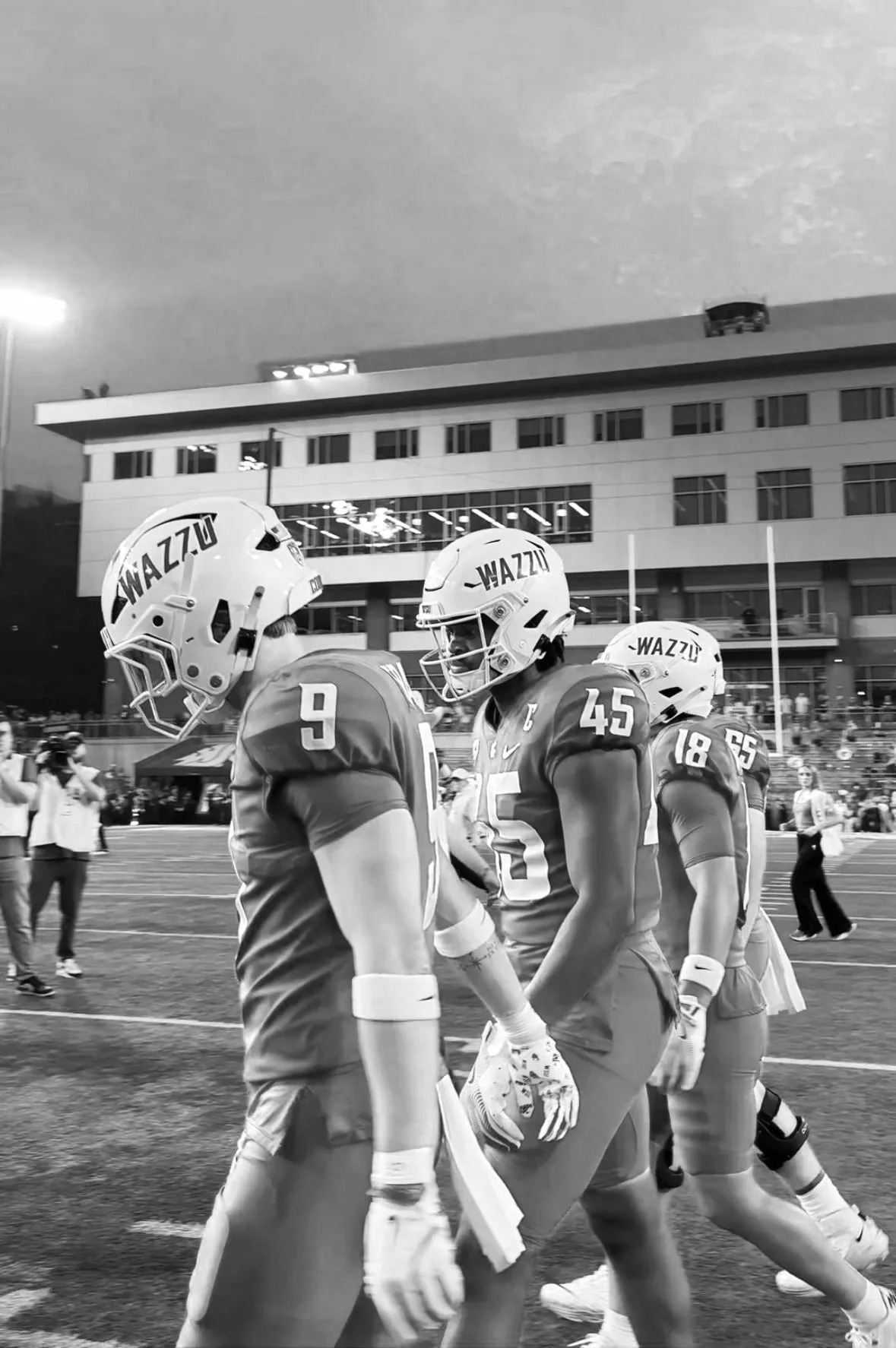 Three football players in uniform walking on the field during a game, wearing helmets with the word "WAZZU" on them.