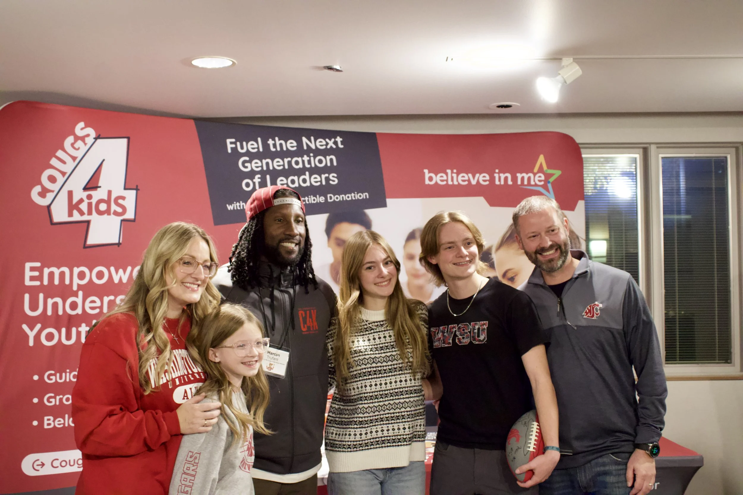 A group of six people, including children and adults, smiling and posing together at an event with a red and white backdrop that reads 'Courage 4 Kids' and 'Empower Youth'. The event promotes leadership and youth support.