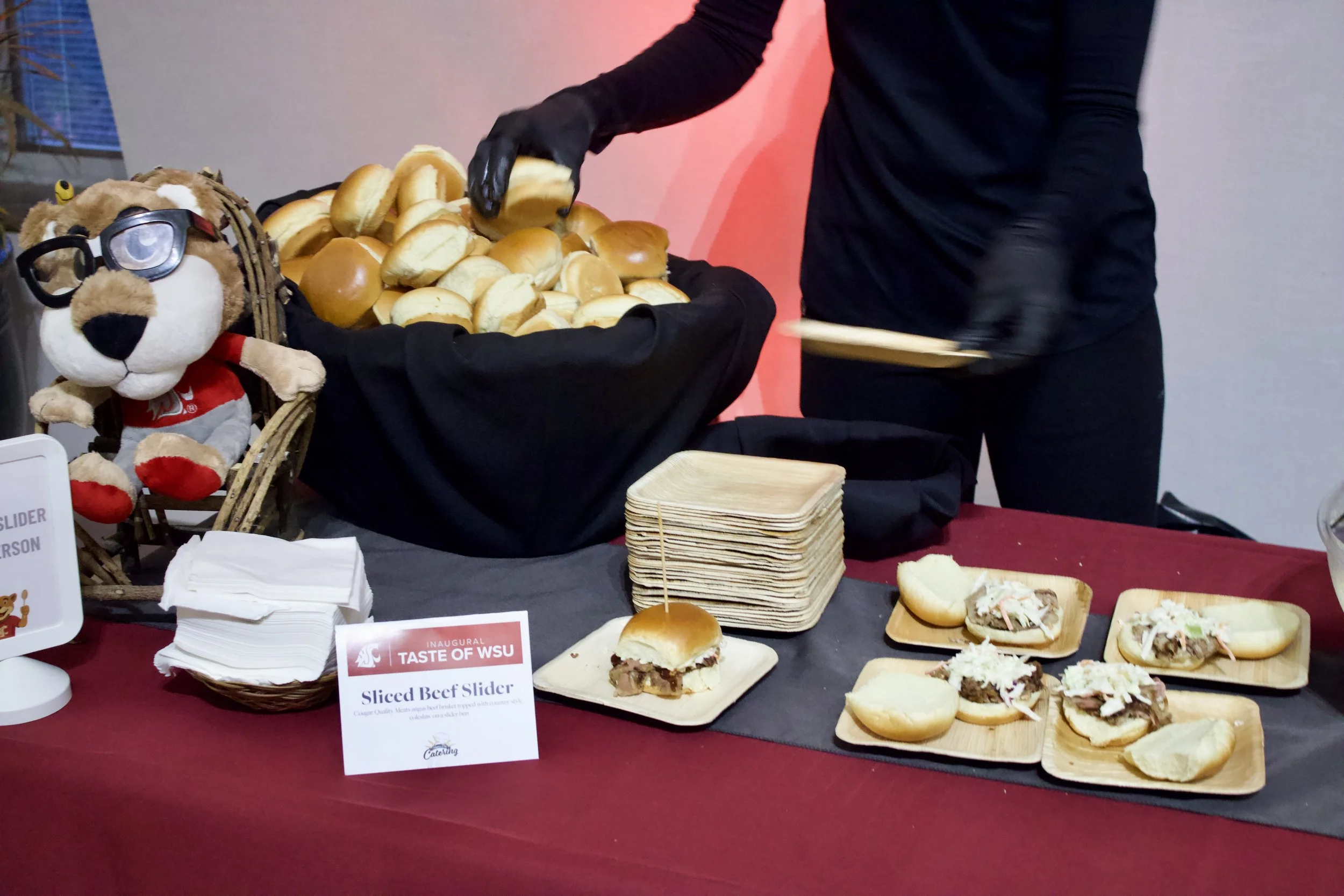 A person serving sliced beef sliders at a food booth with plates of sliders on the table. The table has a stack of wooden plates, napkins, and a sign that reads 'Sliced Beef Slider'. There is a stuffed animal wearing glasses and a Washington State Un