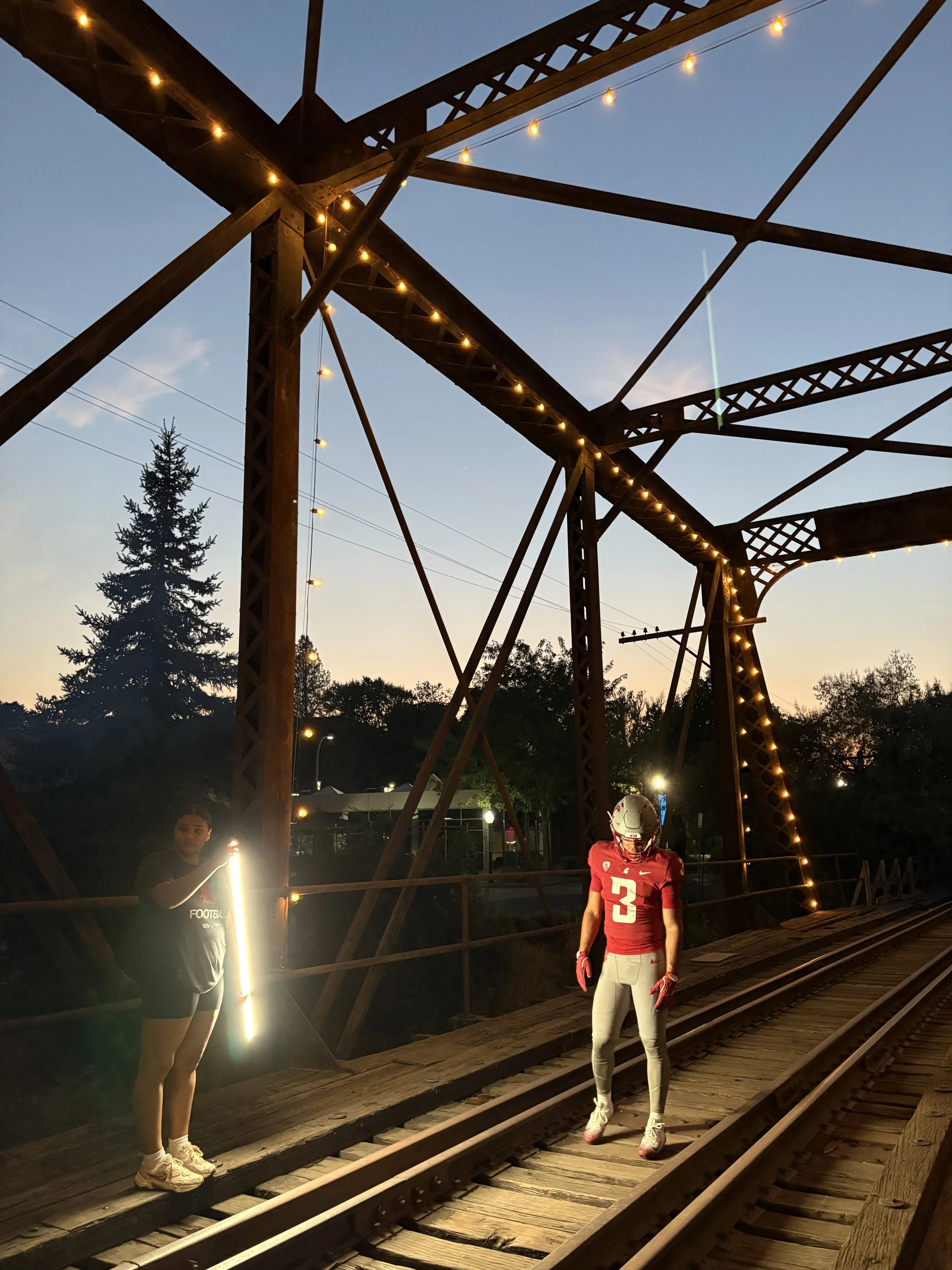 A person in a football uniform with a helmet and gloves walking on a railroad bridge at dusk, with a woman nearby holding a lit phone.
