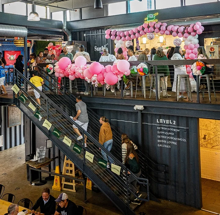 People standing in line at a food counter decorated with pink balloons in a modern indoor setting with a staircase leading to an upper level.