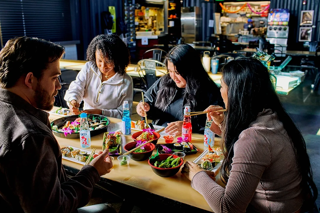 Four people sitting at a sushi restaurant table, enjoying a meal with various bowls of sushi, sashimi, and drinks, in a lively indoor setting.