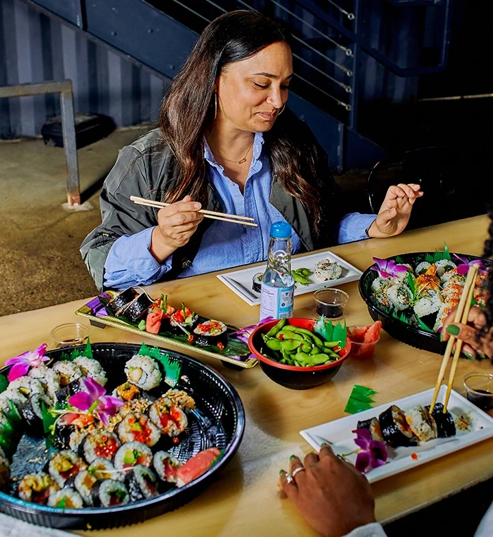 Woman eating sushi at a table filled with various sushi dishes, edamame, and soy sauce, in a restaurant setting.