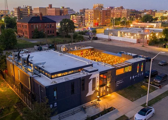 A converted shipping container building with illuminated string lights on a city street at dusk.