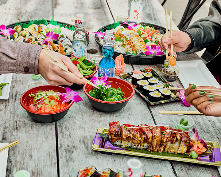A diverse sushi platter with various rolls, sashimi, and garnishes, along with bowls of salad and dipping sauces, on a rustic wooden table where people are using chopsticks to eat.