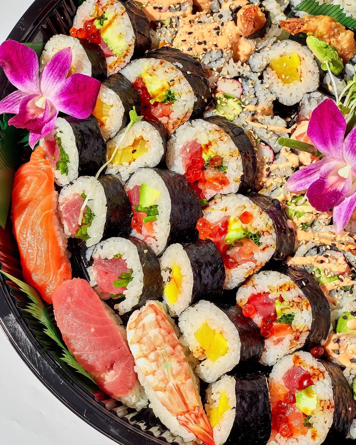 Assorted sushi rolls with salmon, shrimp, tuna, avocado, cucumber, and decorative pink flowers on a black platter.