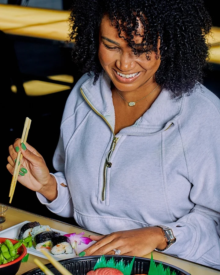 A woman with curly hair smiling, holding chopsticks, sitting at a table with sushi and other food items.