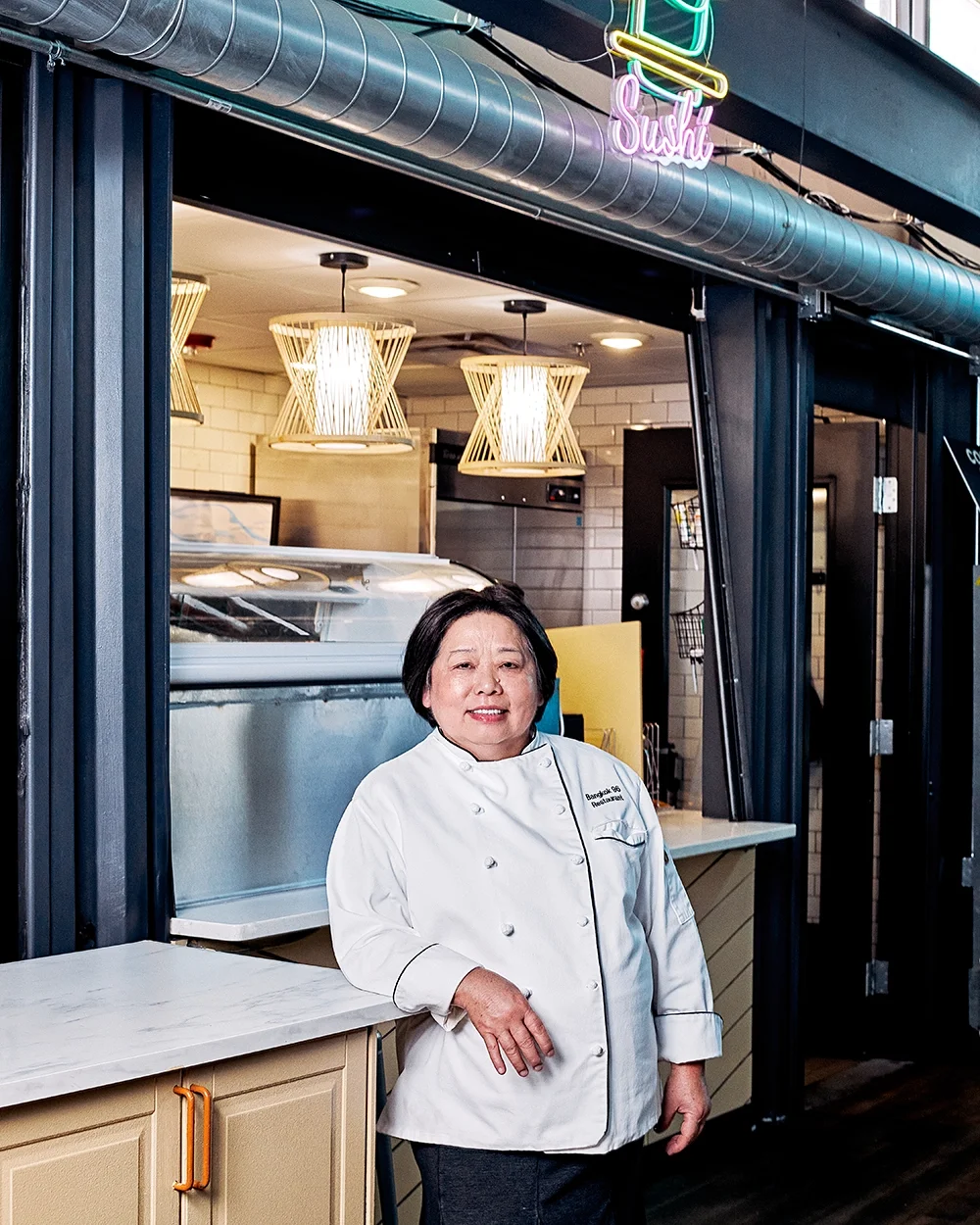 A woman in a white chef's coat standing inside a restaurant, smiling at the camera.