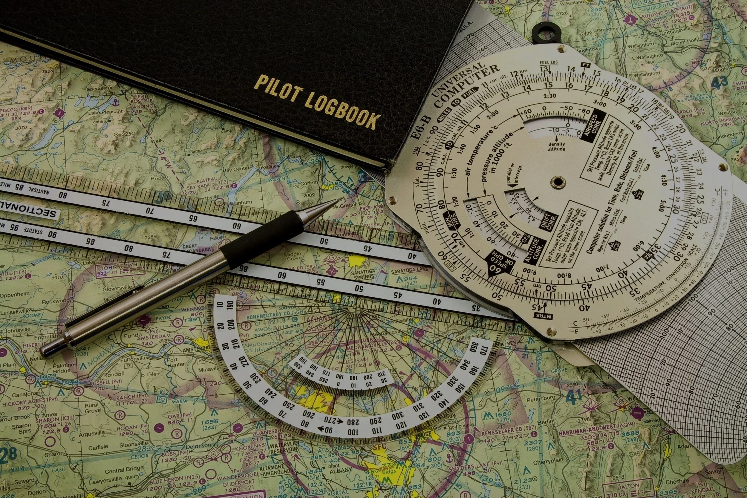 A pilot logbook, a pen, a circular slide rule for calculations, and a detailed sectional map of a geographical area with airports and landmarks.