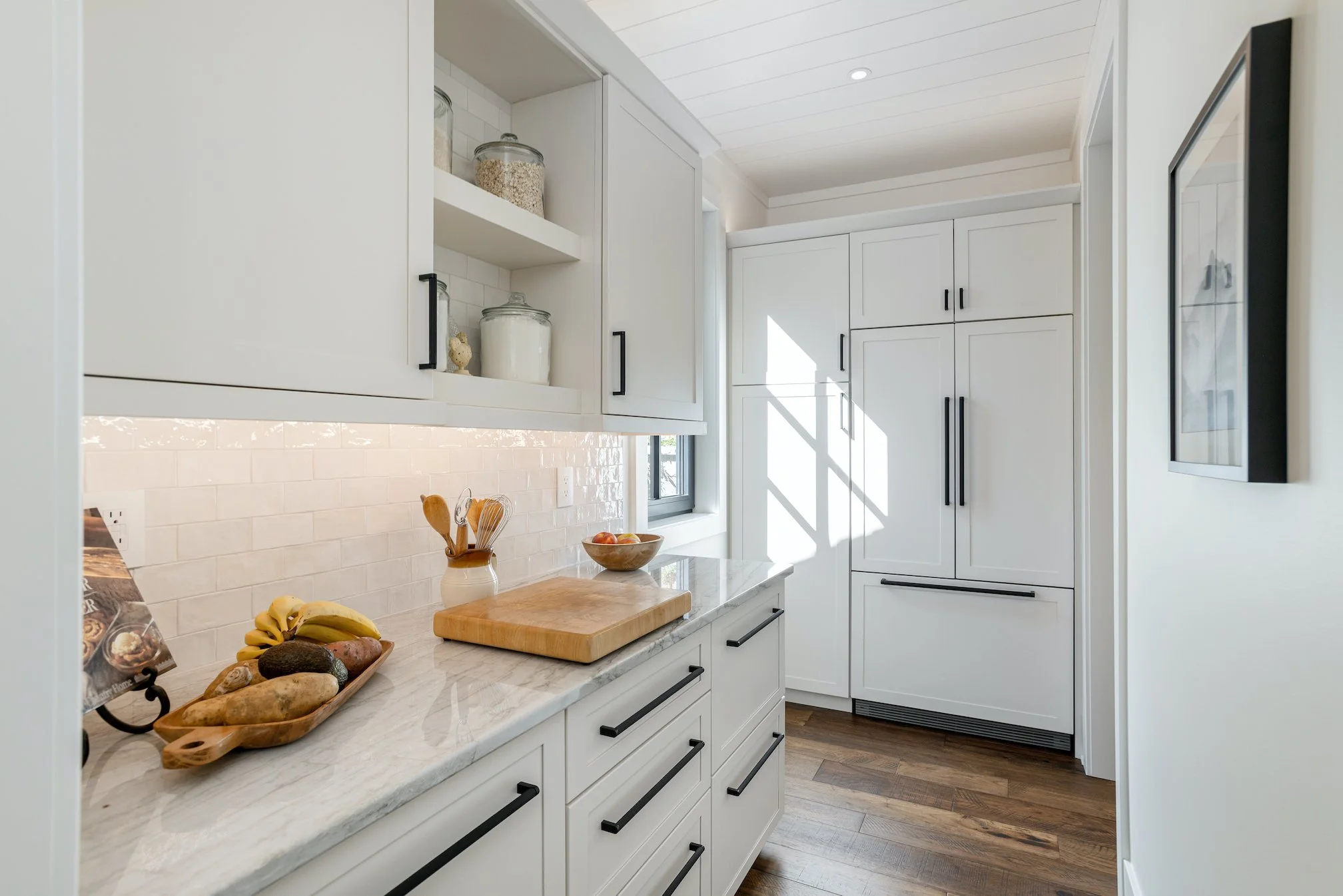 Modern white kitchen with black handles, marble countertop, wooden floor, sunlight through window, open shelving with jars, framed picture on wall.