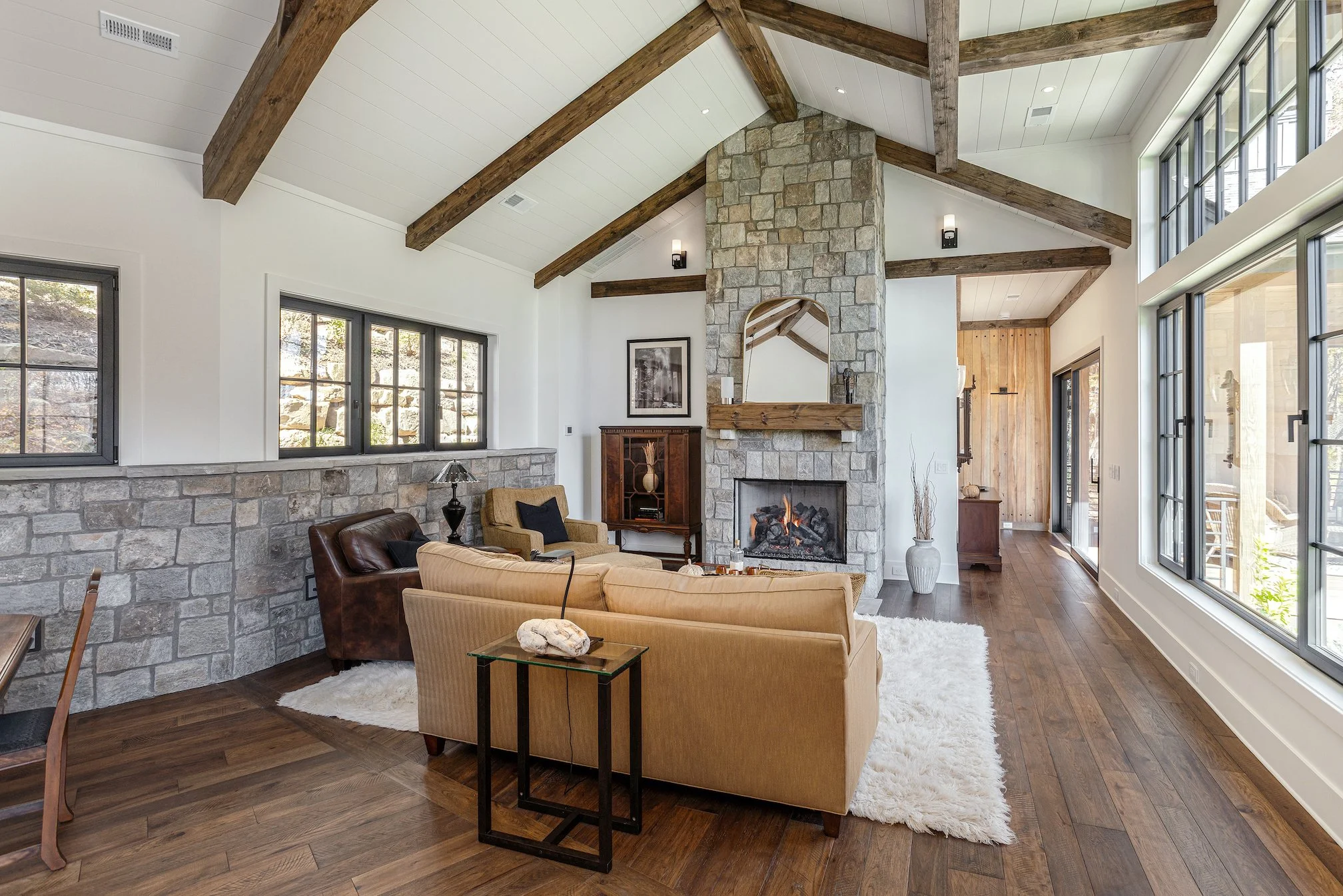 Living room with a stone fireplace, tan and brown sofas, and large windows with black frames, wooden ceiling beams, hardwood floors, and natural light.