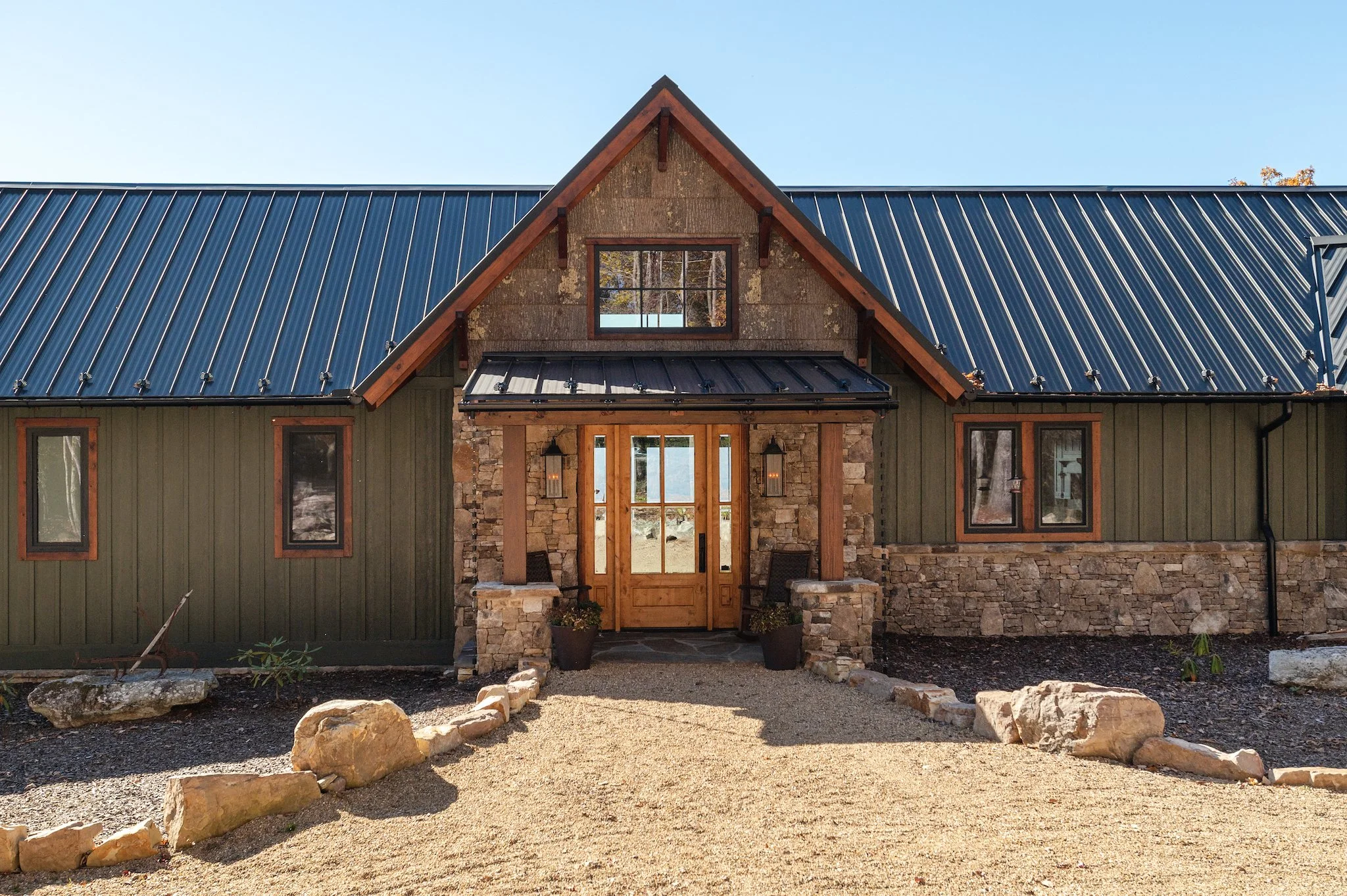 Front view of a modern rustic house with stone and wood exterior, metal roof, and a centered wooden front door with glass panels, surrounded by rocks and gravel landscaping.