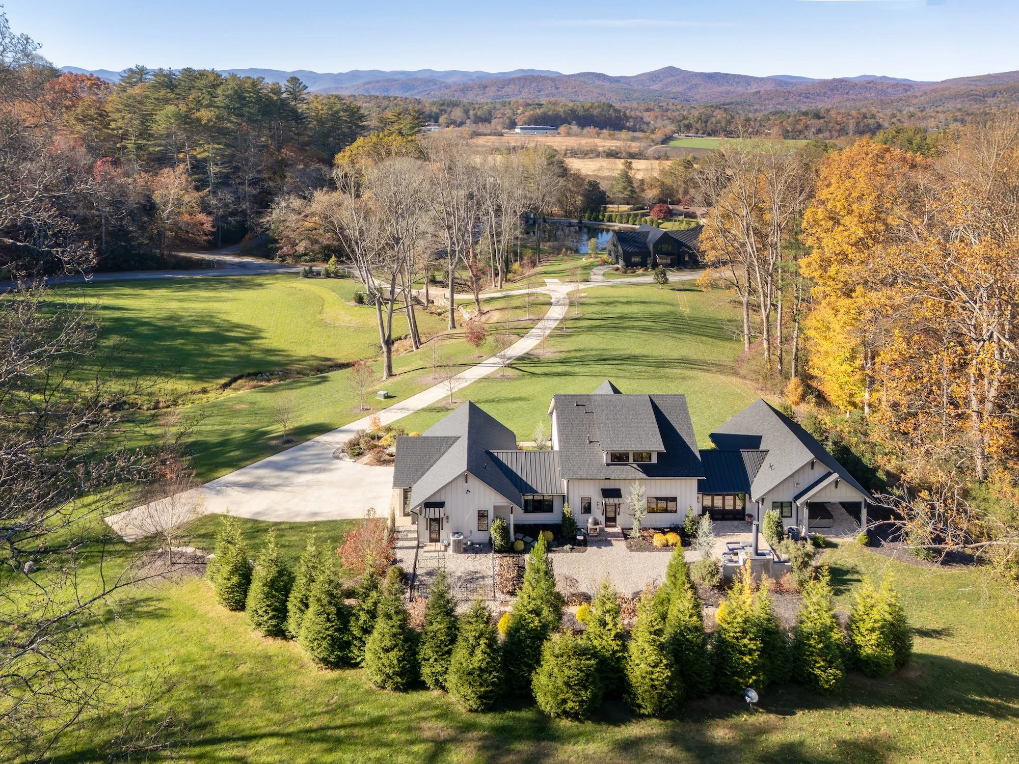 A modern house with a black roof and white exterior, surrounded by greenery and trees, with a gravel path leading to the house, and a large lawn with fall foliage and mountains in the background.