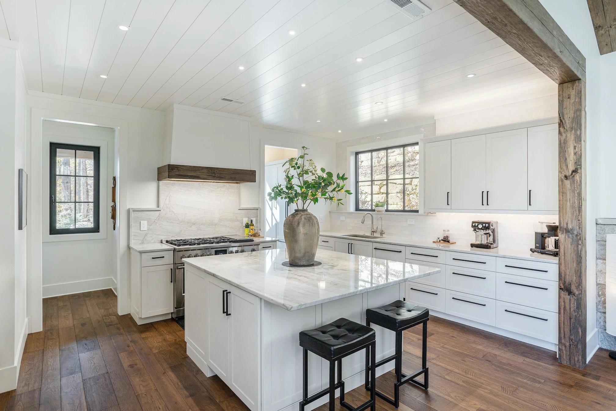 Modern kitchen with white cabinets, marble countertops, a large kitchen island with a vase of green leaves, wooden accents, and black bar stools, illuminated by recessed ceiling lights.