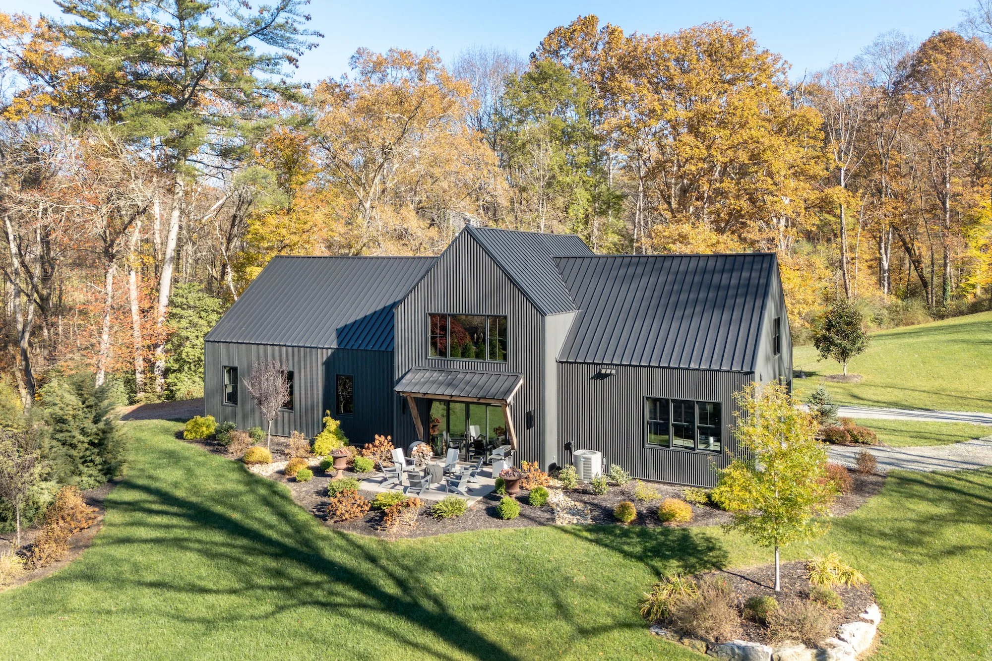 A modern black house with a metal roof surrounded by well-manicured green lawn and landscaping, with colorful trees in the background, during autumn.