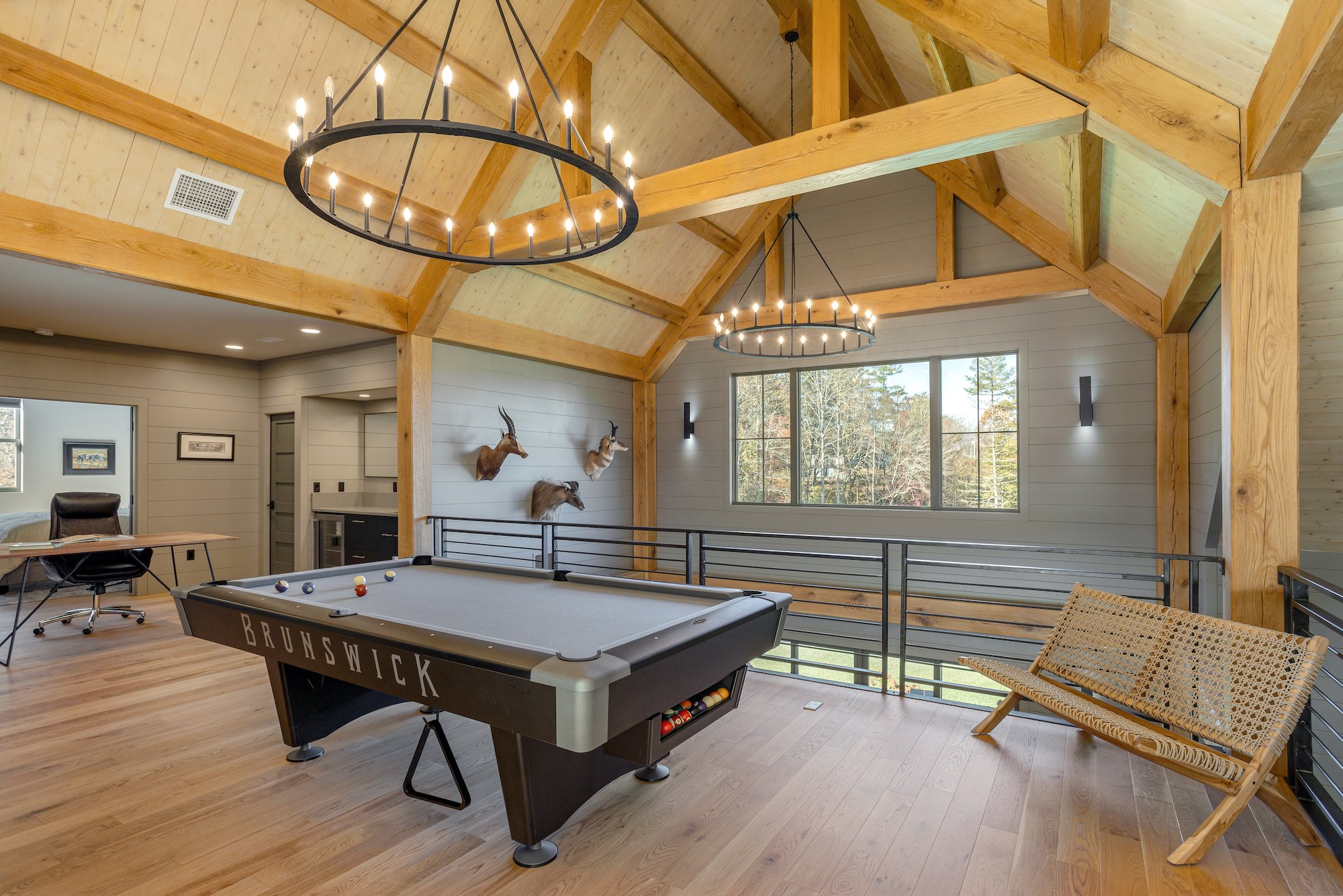 Interior of a spacious game room with wooden beams and paneling, featuring a Brunswick pool table, wall-mounted animal heads, large window, chandelier lights, a woven bench, and a desk with a black chair.