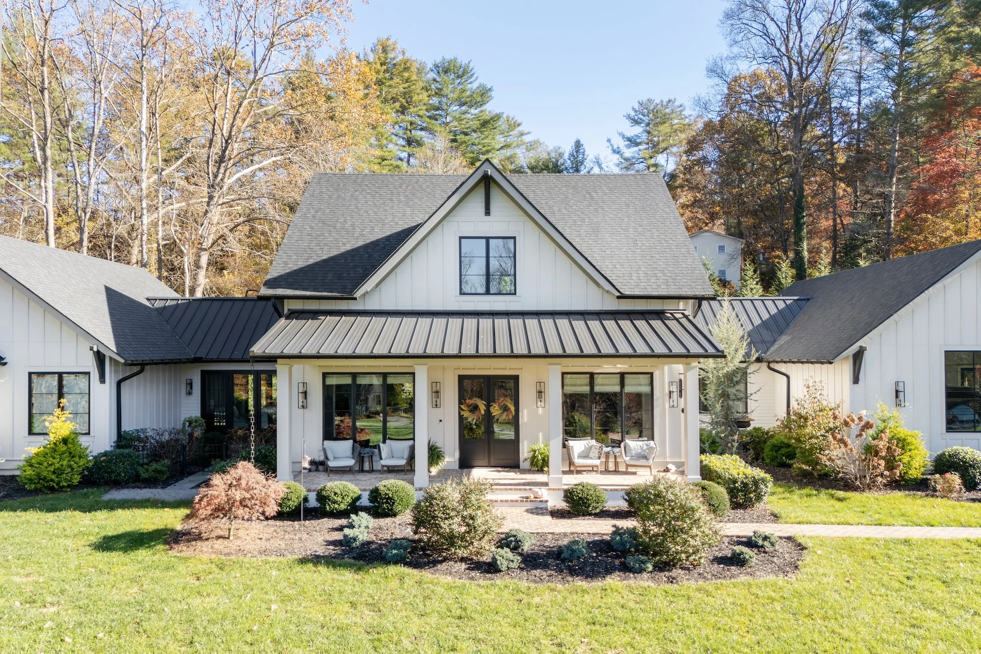 Front view of a modern white house with a gray gabled roof, black window frames, and a porch with outdoor furniture, surrounded by a landscaped garden with bushes and trees, and autumn-colored forest in the background.