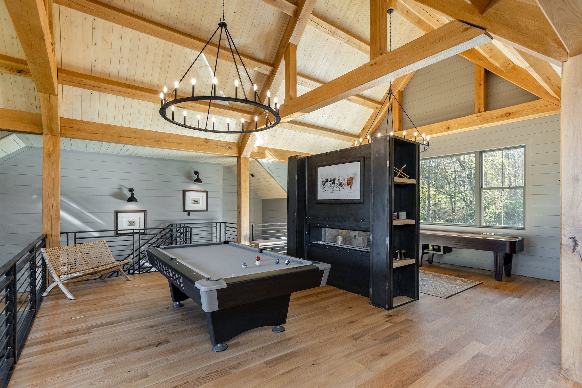 Interior of a loft with wooden beams, a chandelier, a pool table, a black bookshelf with artwork, a window, and a shuffleboard table.