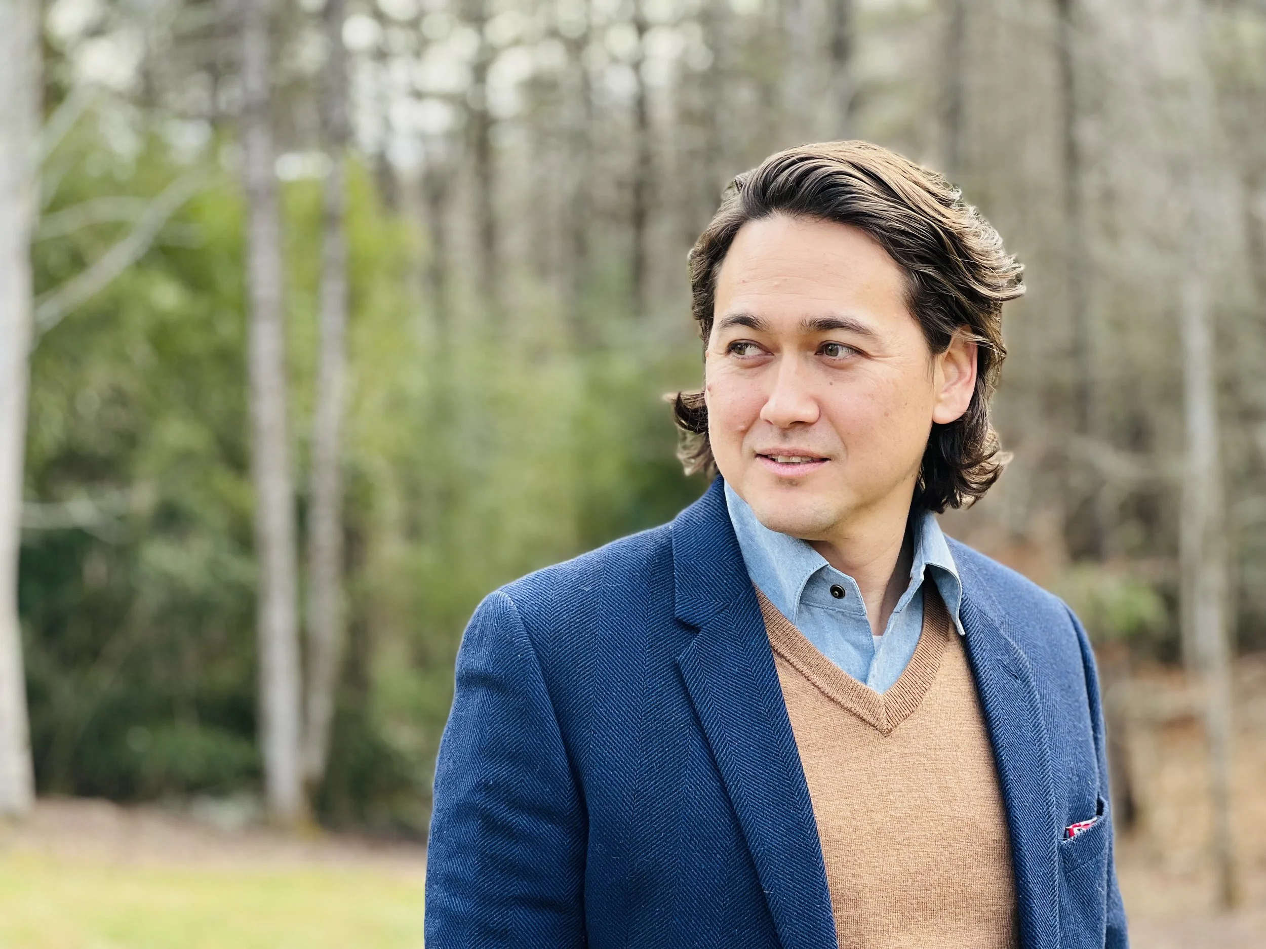A well-dressed man with dark brown, shoulder-length hair stands outdoors in a wooded area. He is wearing a blue blazer, light blue shirt, and a tan sweater vest. He is looking to his right with a subtle smile, surrounded by trees with a background of blurred greenery.