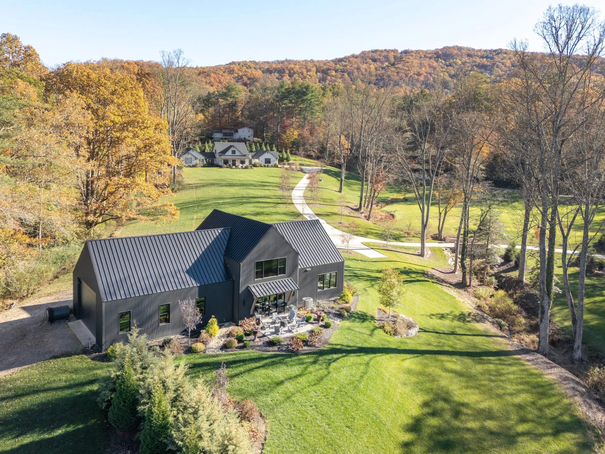 Aerial view of a modern black house with a metal roof, surrounded by green lawn and trees, in a hilly area with some other houses in the background, and mountains in the distance during fall.