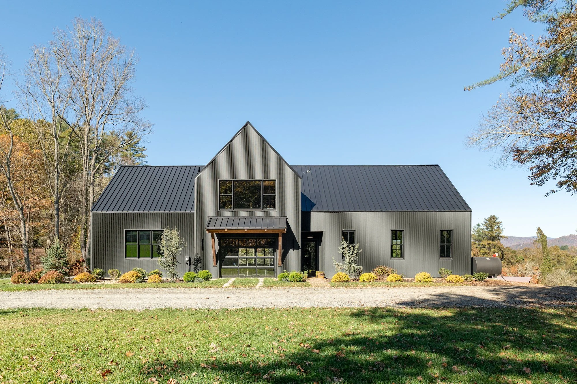 A modern two-story house with a black metal exterior, set on a grassy lawn with shrubs and trees, under a clear blue sky.