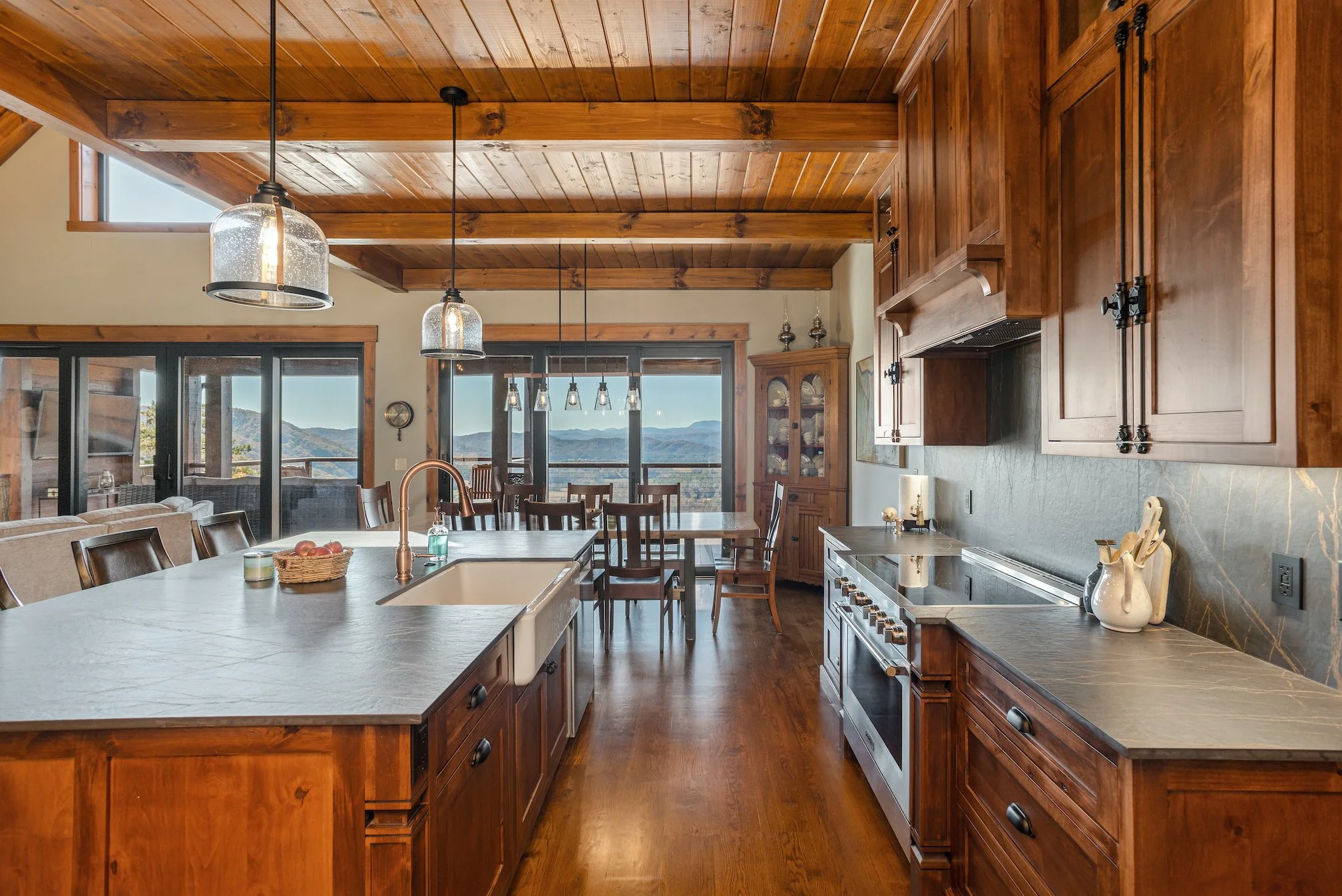 A spacious kitchen with wooden cabinets and ceiling, a large island with a sink, hanging pendant lights, and a dining area with chairs near large windows showing mountain views.