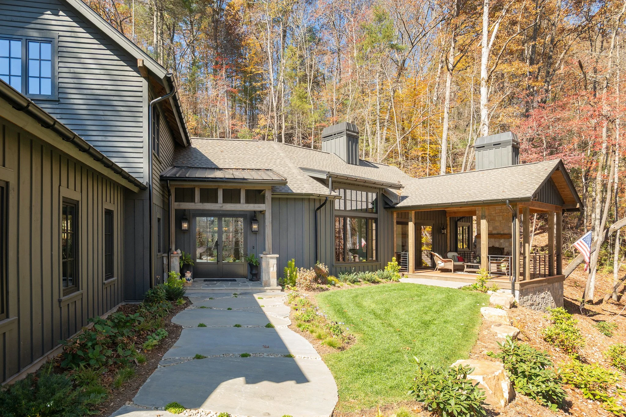 Front view of a modern house with a stone porch and lawn, surrounded by trees with autumn leaves