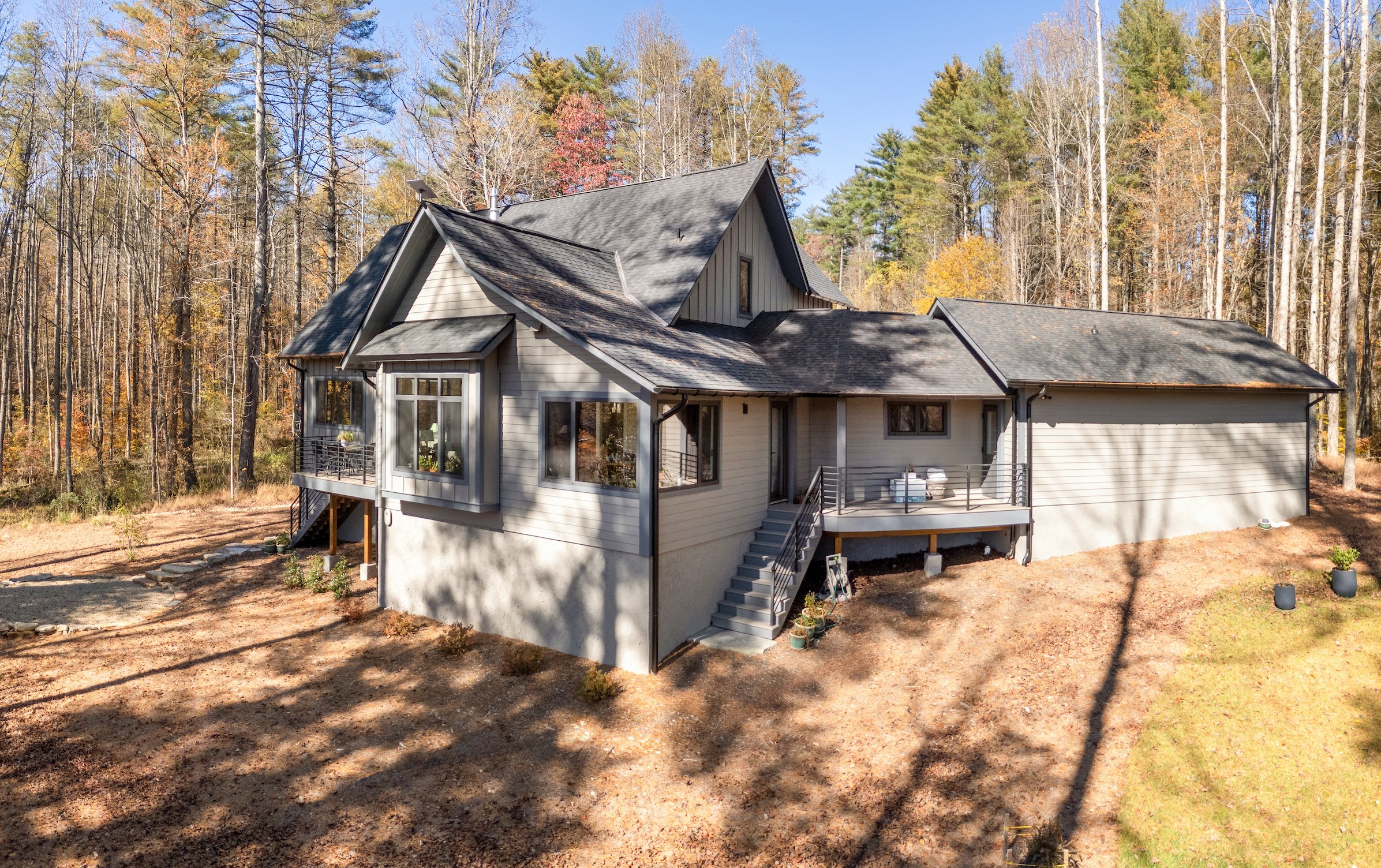 A modern house surrounded by trees in autumn, with a deck, stairs, and a part of a backyard visible.
