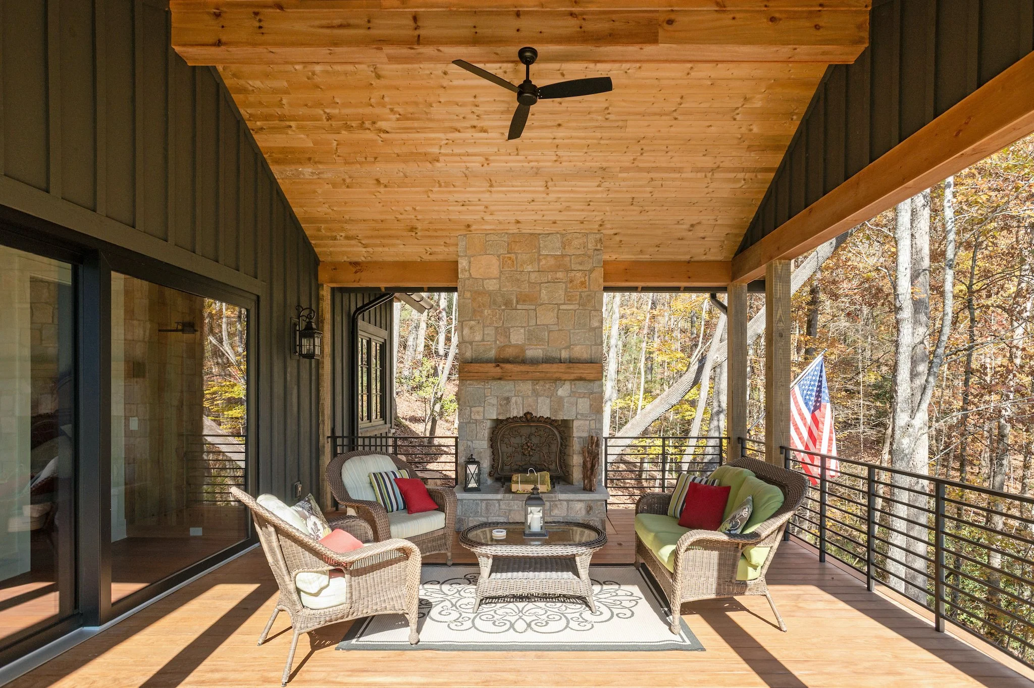 Outdoor covered porch with wicker furniture, a fireplace with a stone chimney, and a ceiling fan, overlooking a wooded area with trees and an American flag.