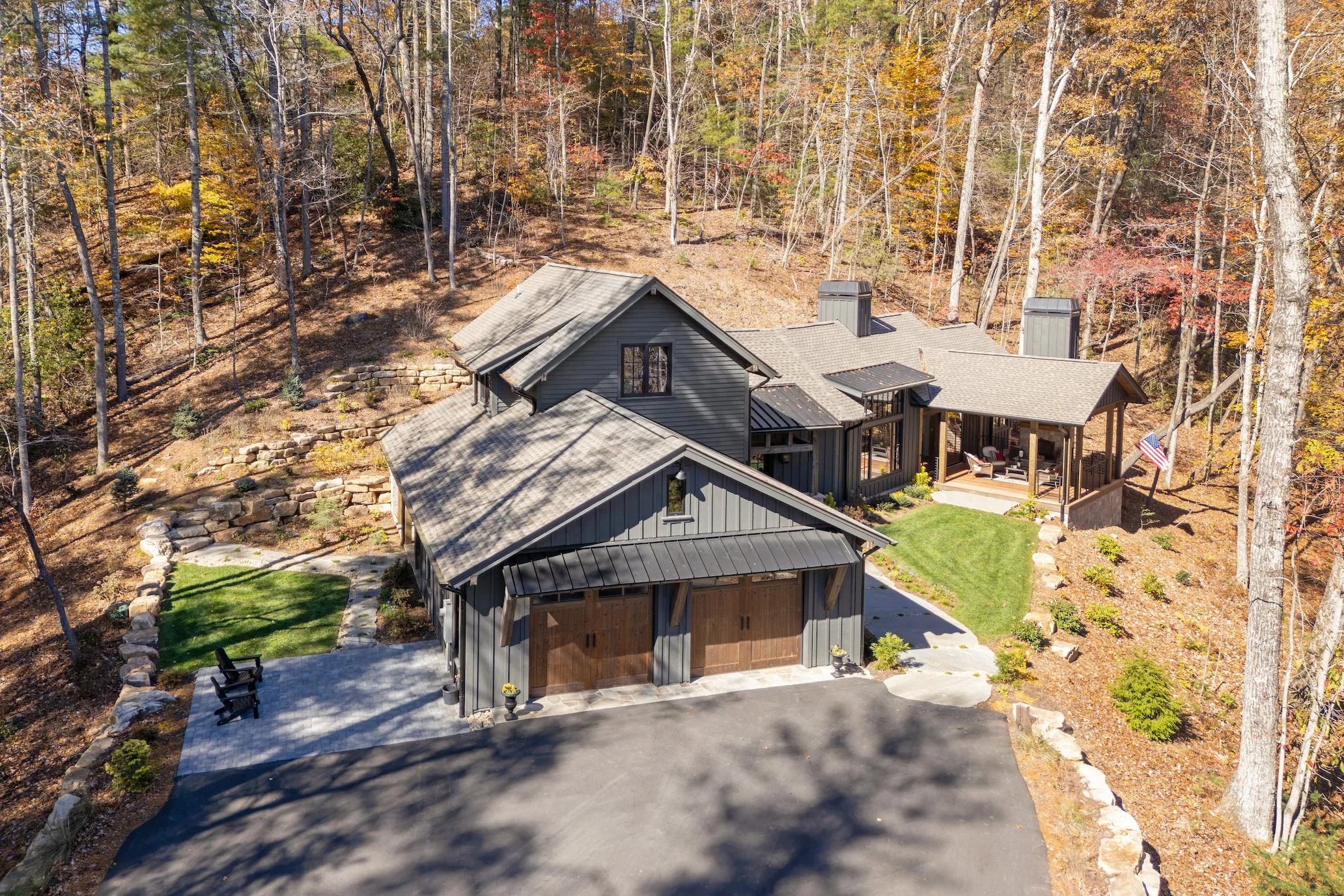 A large modern house with gray siding and multiple gabled roofs surrounded by autumn trees. The house features a front porch with seating, a manicured lawn, and a paved driveway.