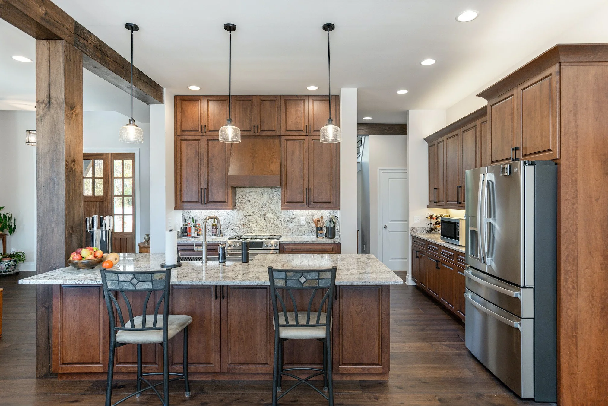 Modern kitchen with wooden cabinets, granite countertops, stainless steel refrigerator, and a kitchen island with two chairs, pendant lighting, and a windowed door in the background.