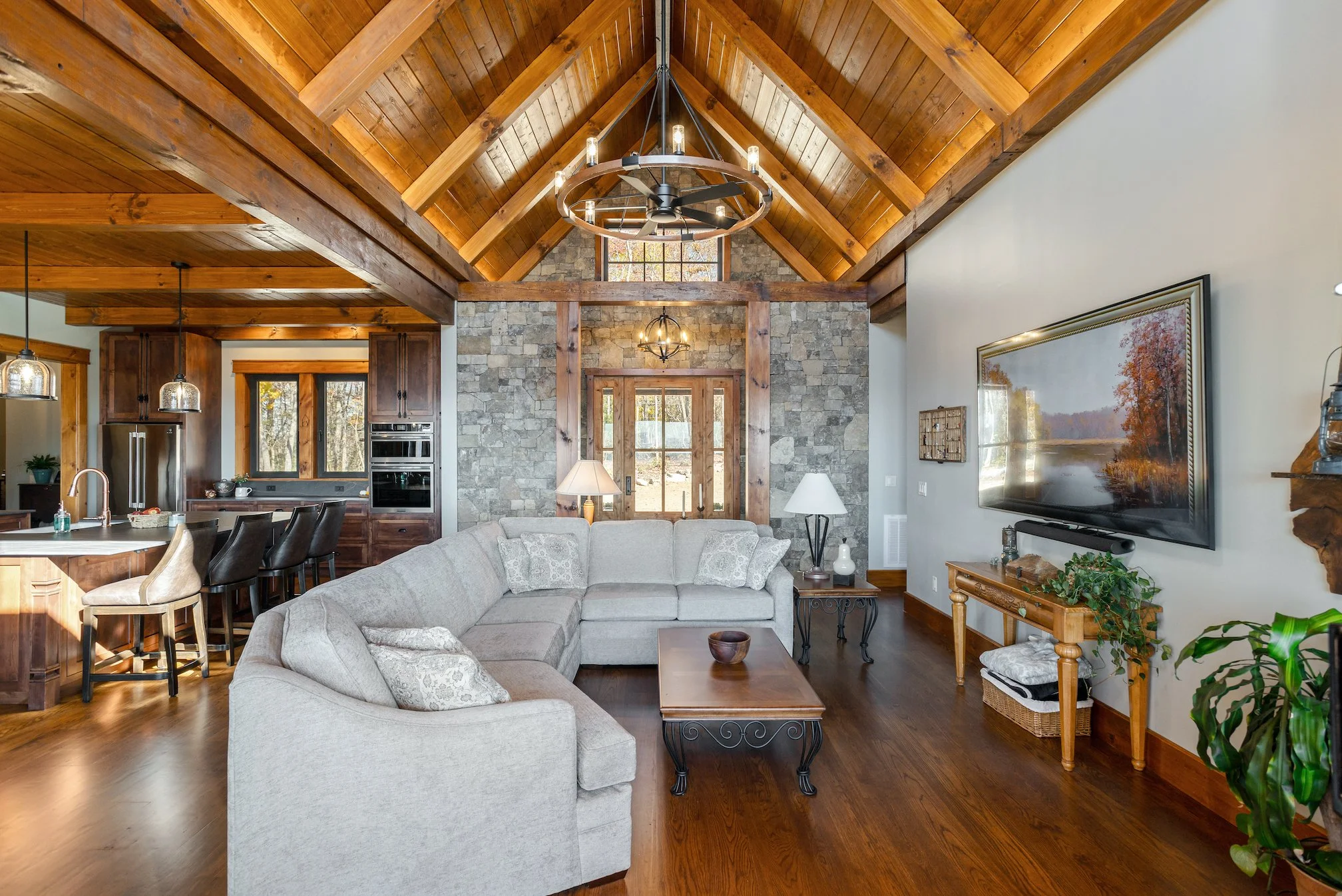 Living room with high wooden vaulted ceiling, stone accent wall at the front, and a large white sectional sofa facing a wall-mounted TV. The room features wooden trim, hardwood floors, side tables with lamps, and decorative plants.