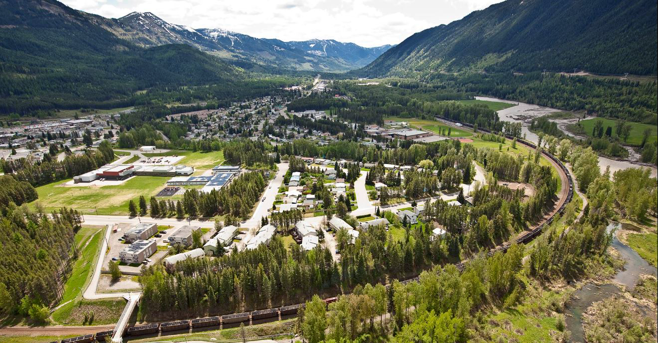 Aerial view of Sparwood, BC in a valley surrounded by green mountains, with a river flowing through the area and a train track on the right side.