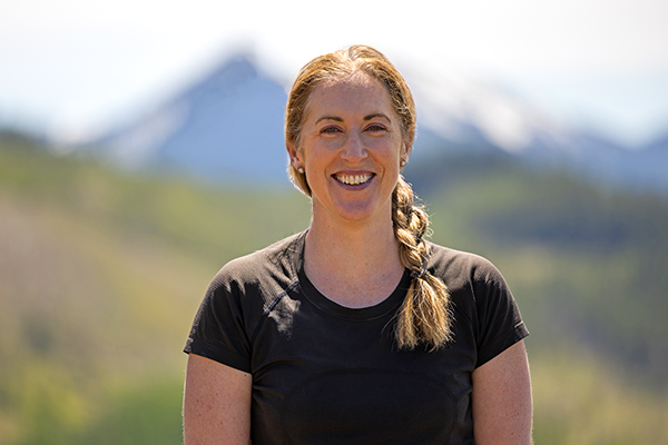 Abby Cousins, General Manager, with long blonde hair wearing a black blazer and white shirt, smiling in an indoor setting with blurred background of shelves and plants.