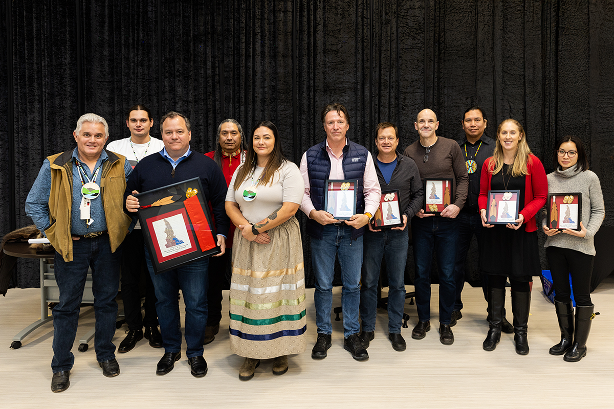Group of ten people standing indoors against black curtains, holding framed artwork and award items, celebrating an achievement.