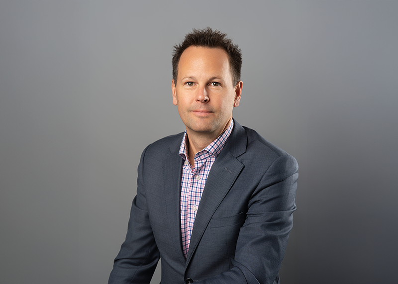 Portrait of David Morgan, Chief Financial Officer, wearing a checked shirt and gray blazer against a plain gray background.