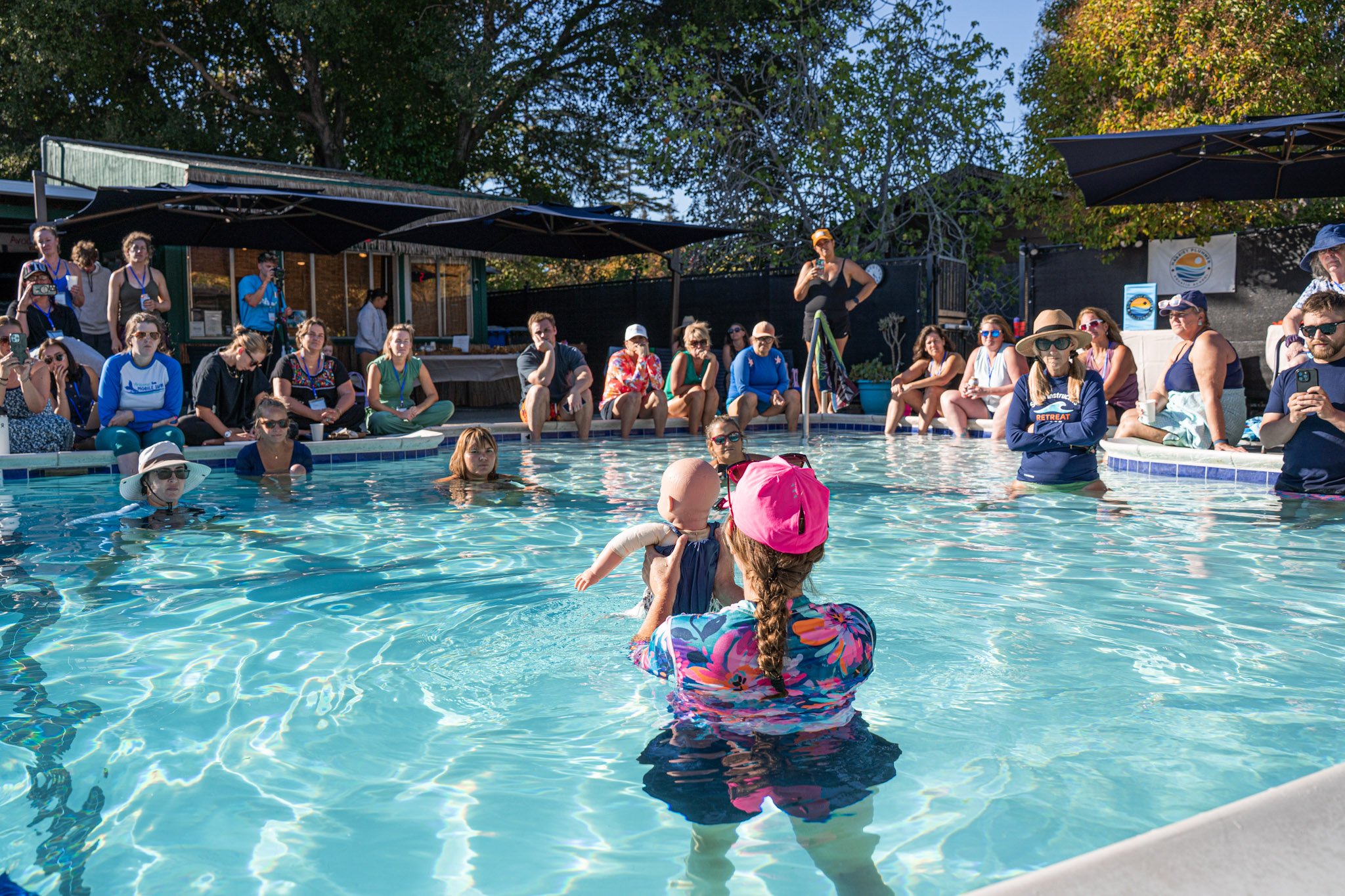 A woman in a pink hat holding a baby in a swimming pool during a poolside event with spectators seated and standing around.