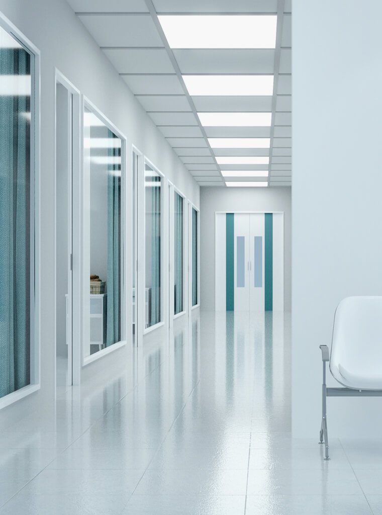 A clean, modern hospital corridor with glass doors on the left and a white chair on the right, leading to an elevator at the end.