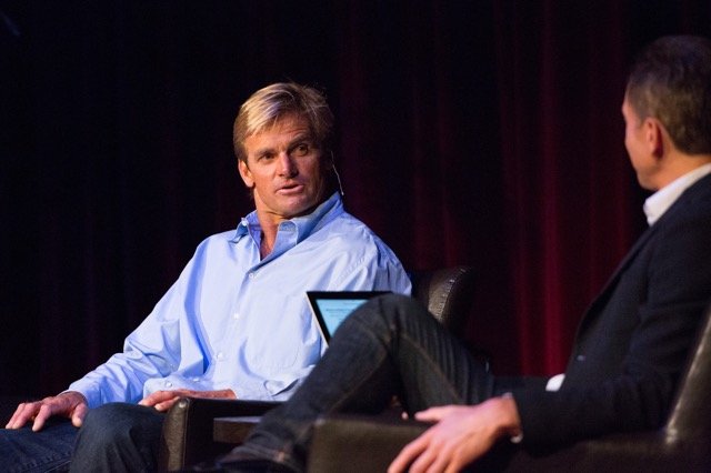 Two men sitting on a stage engaged in a conversation, with a dark curtain in the background.
