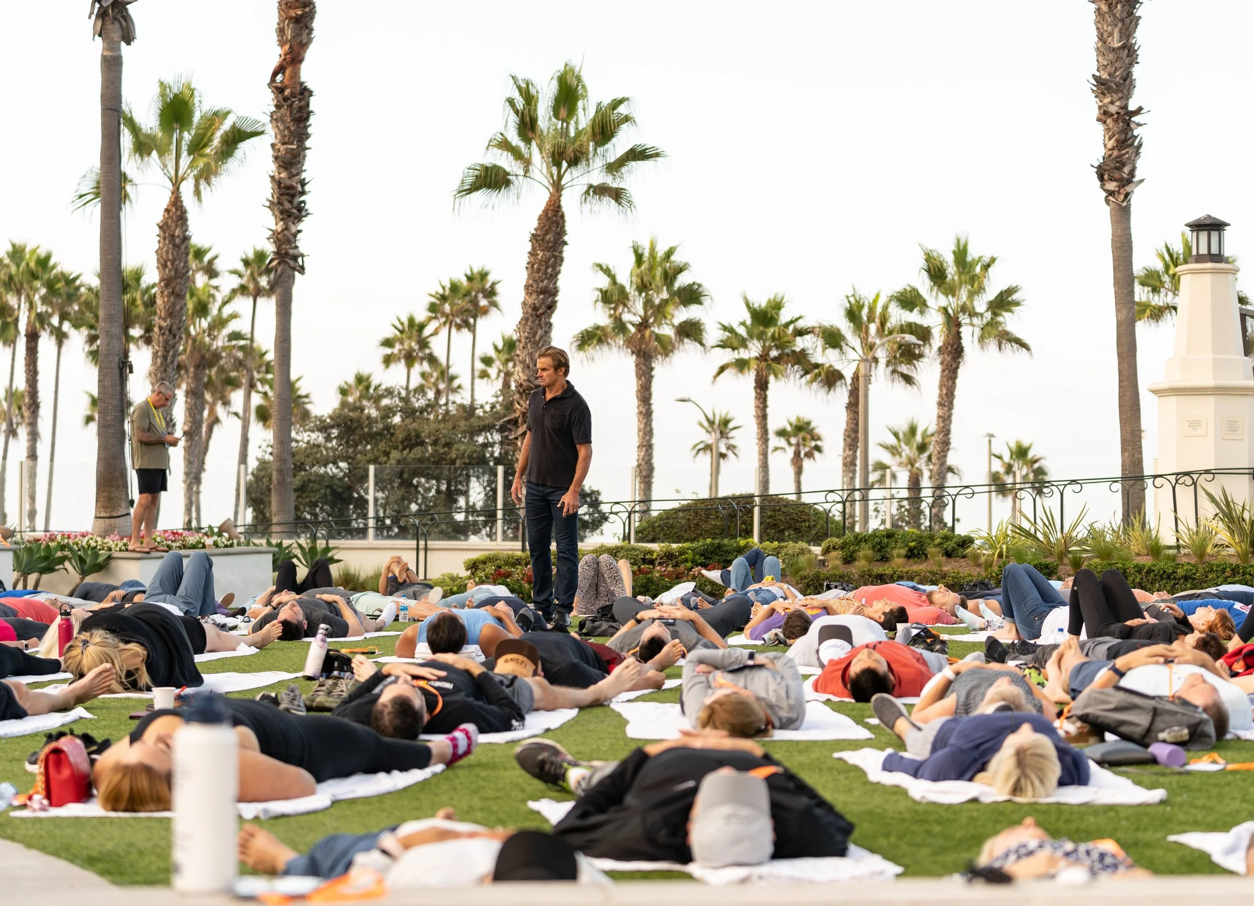 A large group of people participating in an outdoor yoga session on a grassy area, with many lying on yoga mats, surrounded by palm trees and a cloudy sky.