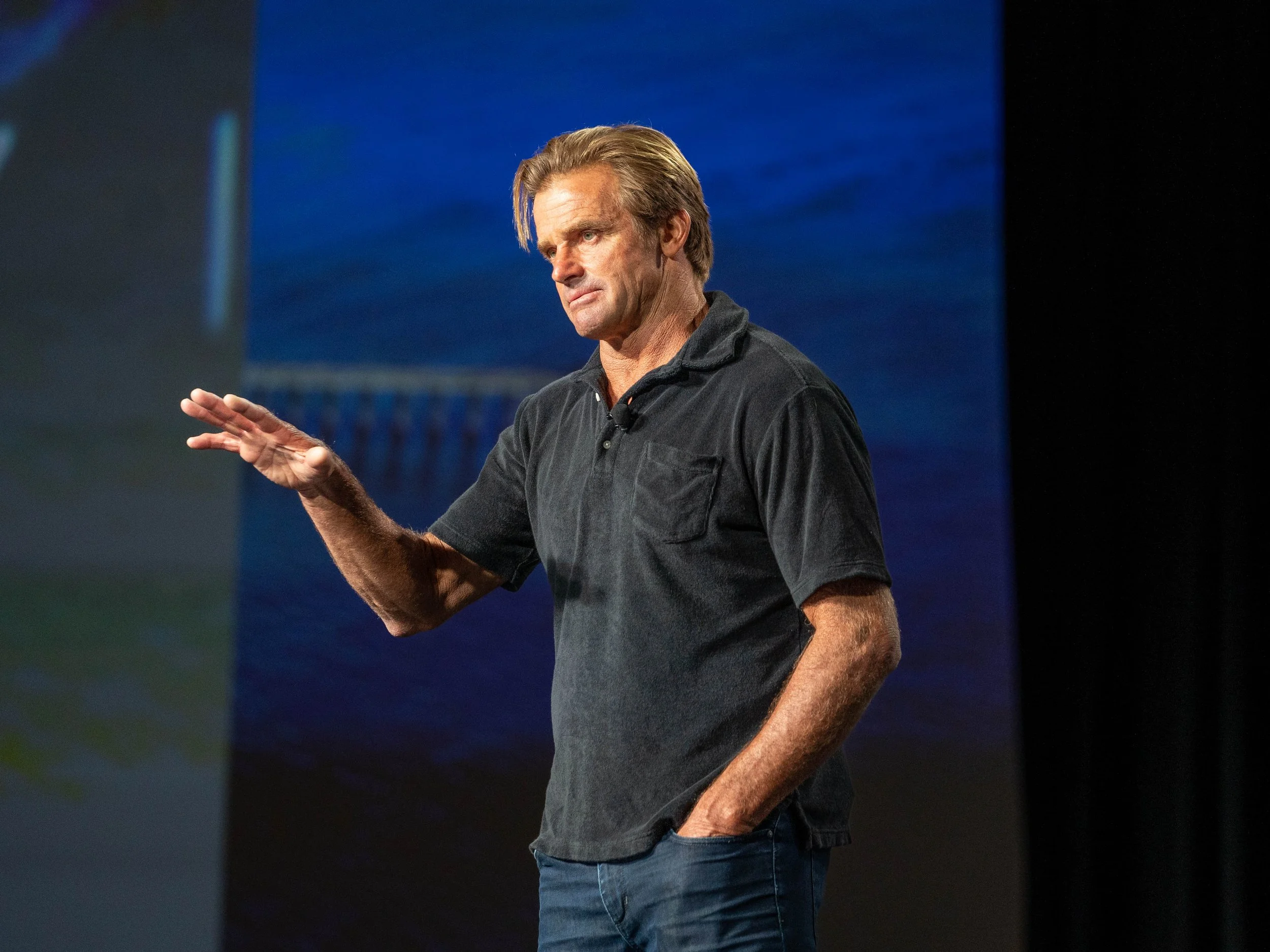 A man with blonde hair, wearing a dark gray polo shirt and blue jeans, is standing on a stage and gesturing with his right hand. The background features a large blue screen.