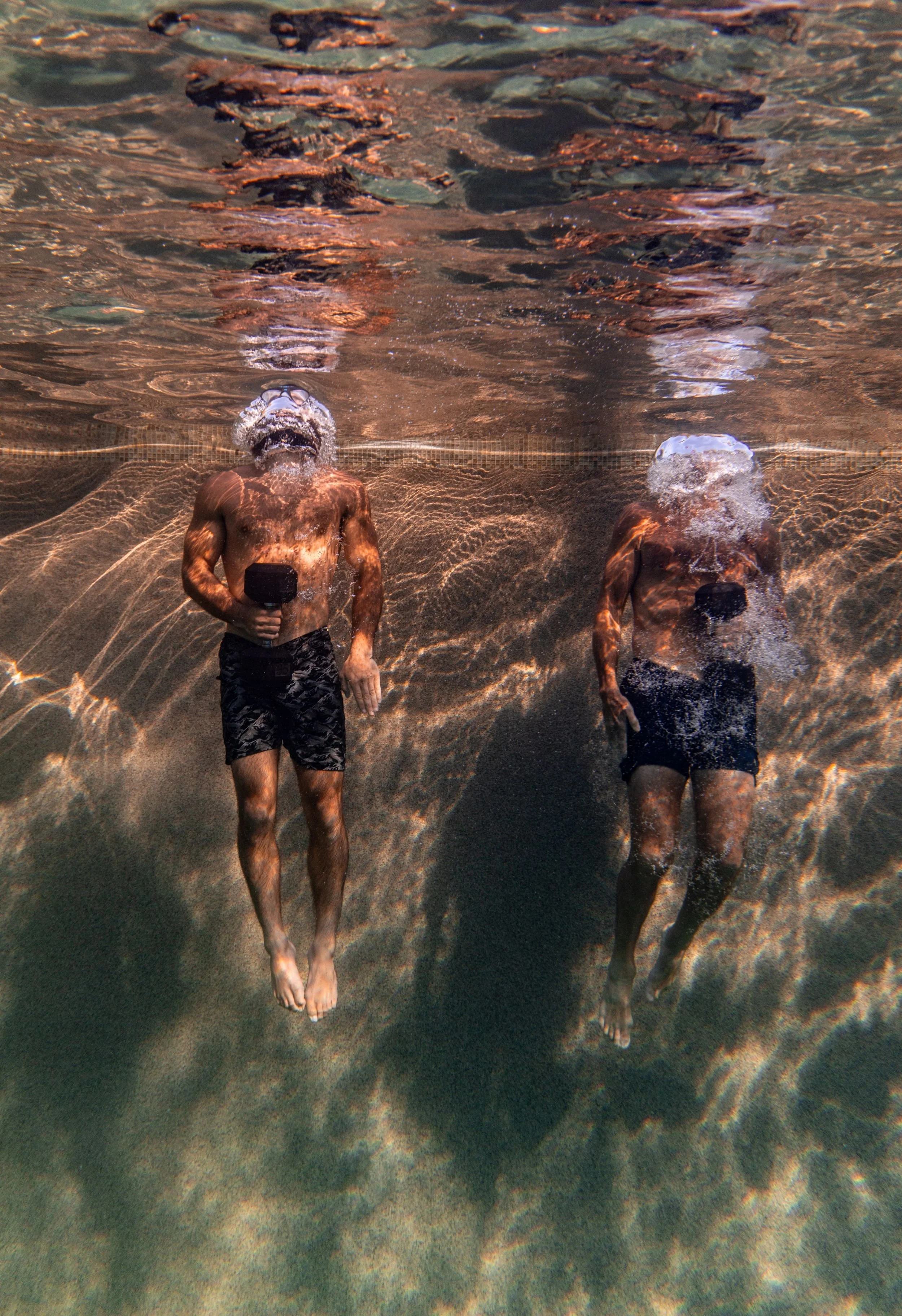 Two men are underwater in a pool or body of water, wearing black swim shorts and transparent goggles. They are holding underwater cameras, and their heads are submerged, creating bubbles and reflections on the water surface.