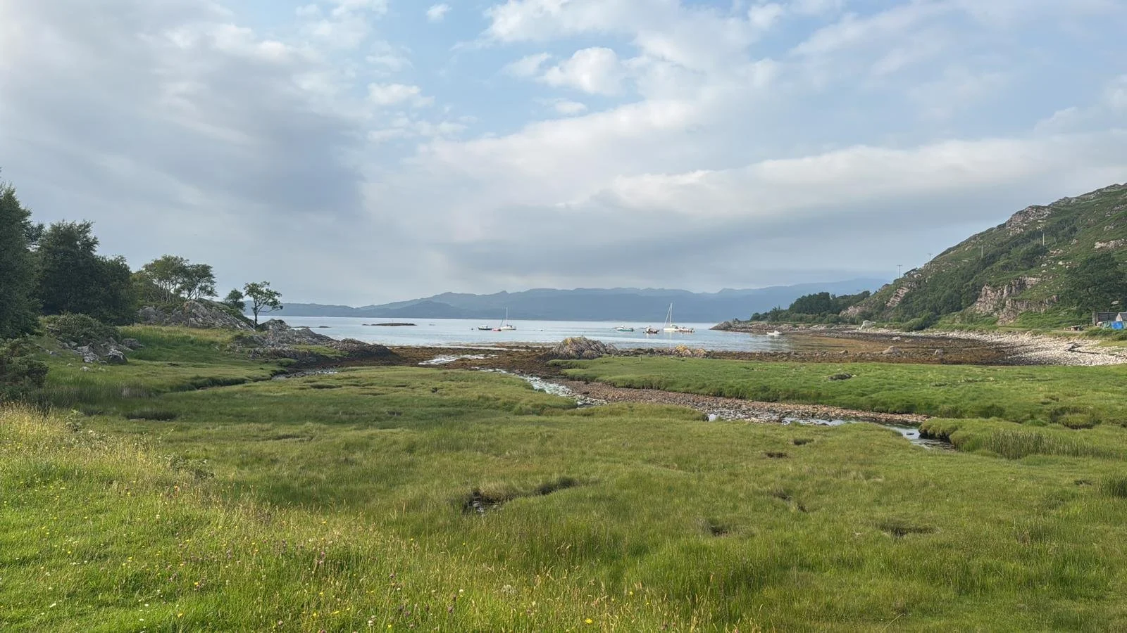 Scenic view of a grassy coastline with small streams flowing into the water, sailboats in the distance, rocky hills on the right, and a partly cloudy sky overhead.